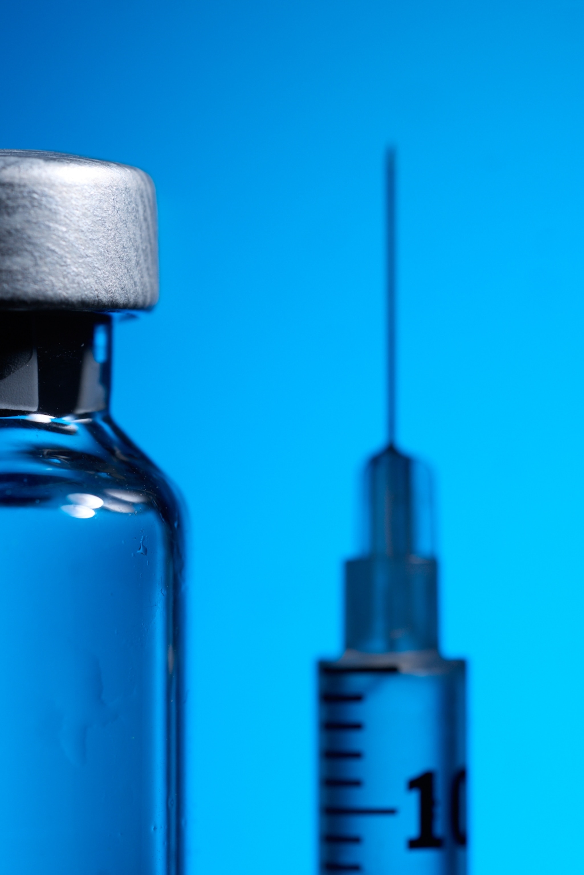 A close-up view of a glass vial in the foreground with a needle and syringe in the background