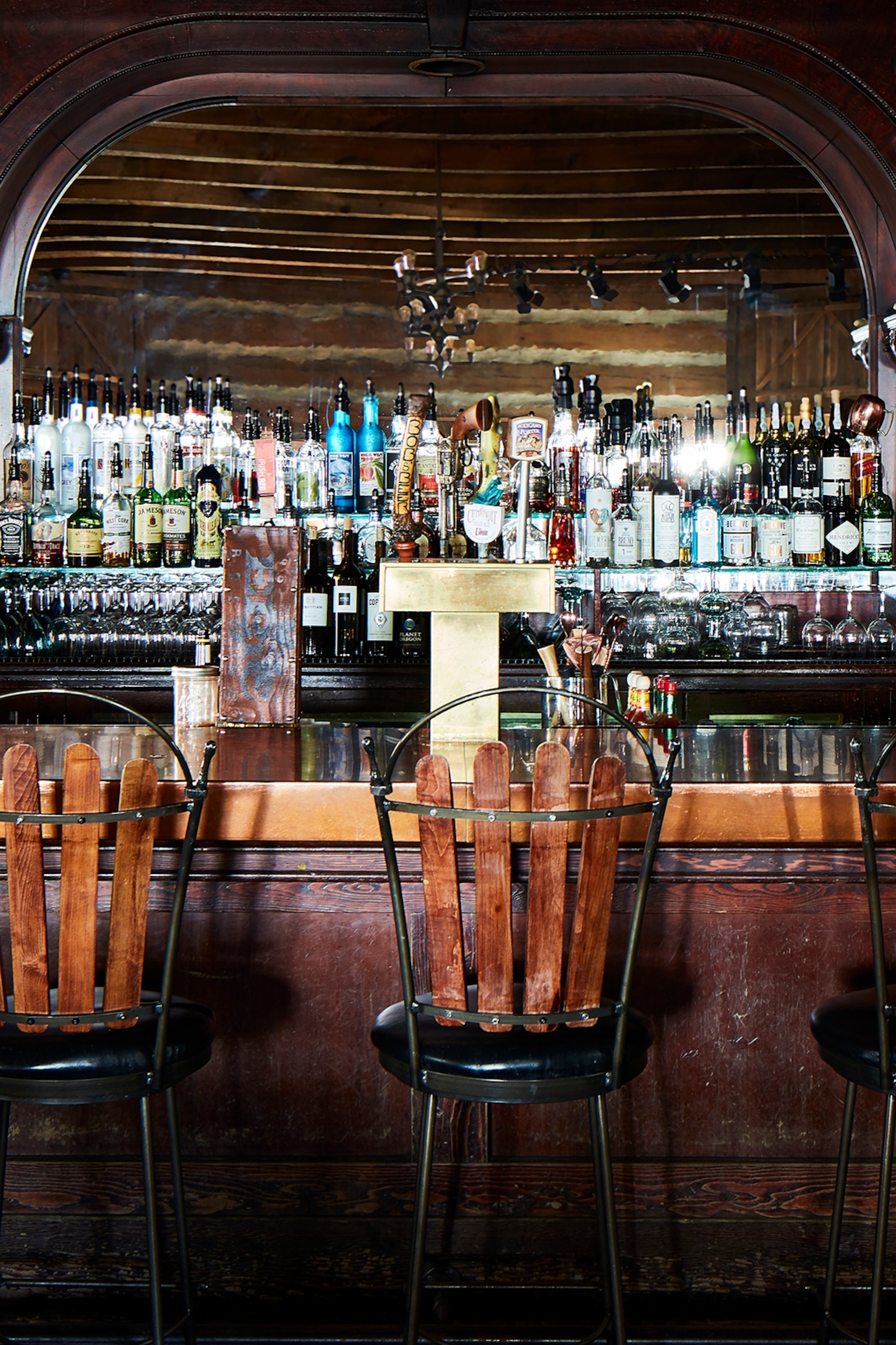 A bar with barrel-back stools and a wooden, arched wall presenting bottles of alcohol and spirits in a spotlight.