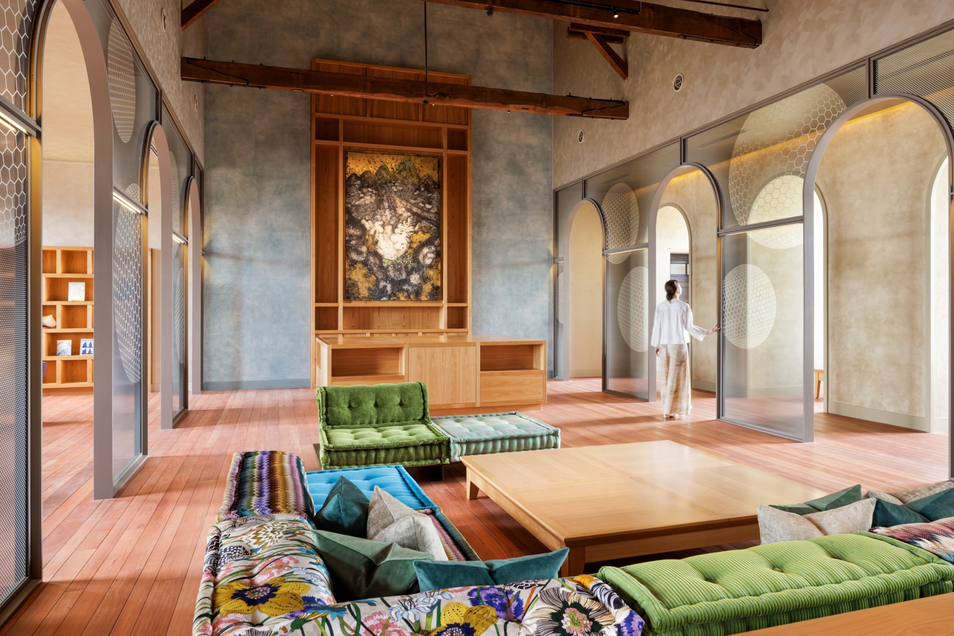 A spacious lounge with wood floors, colorful seating, arched partitions, and a large artwork displayed on the far wall.