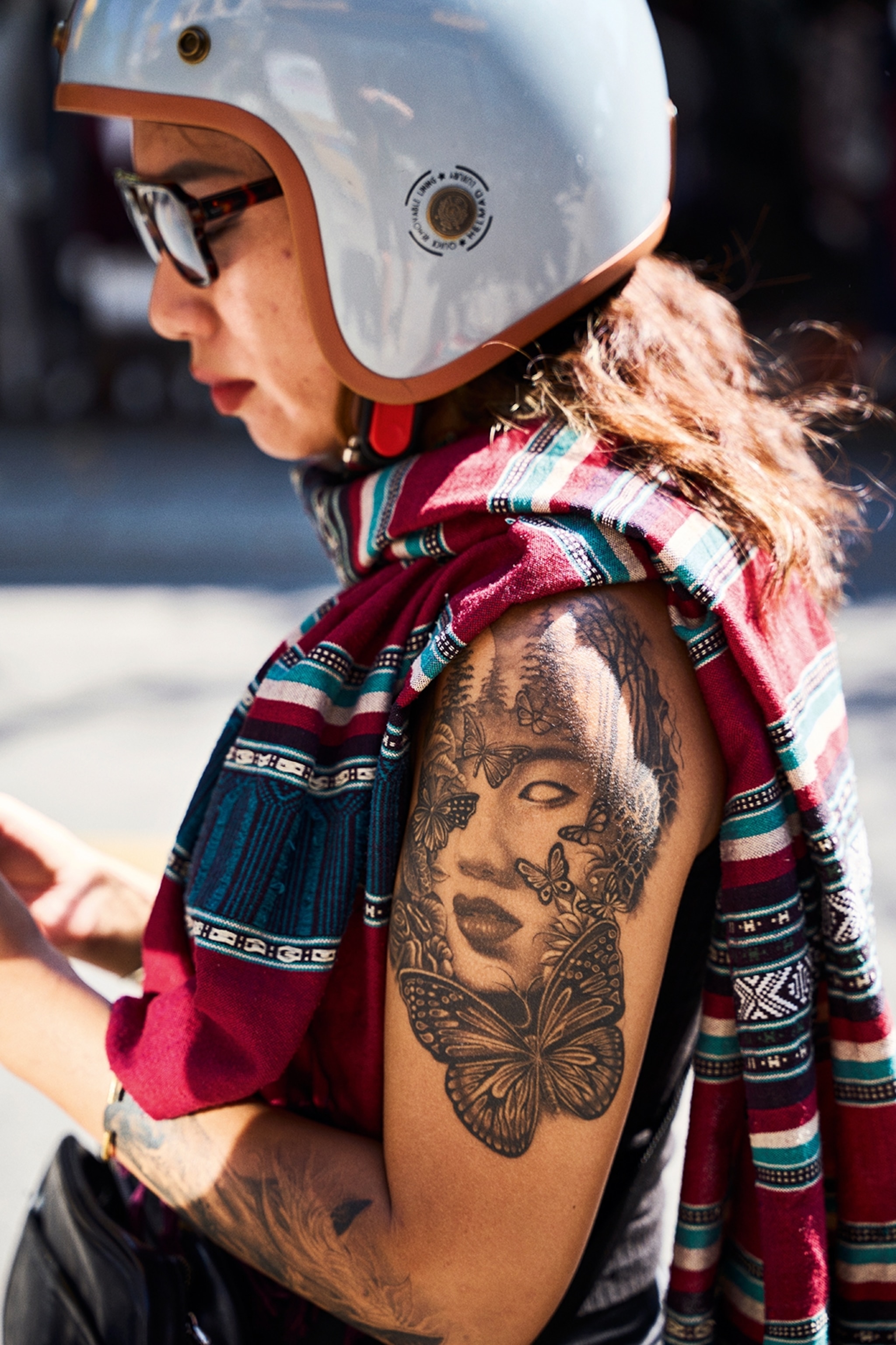 A female biker resting on the side of a street with her helmet on, revealing a shoulder tattoo.
