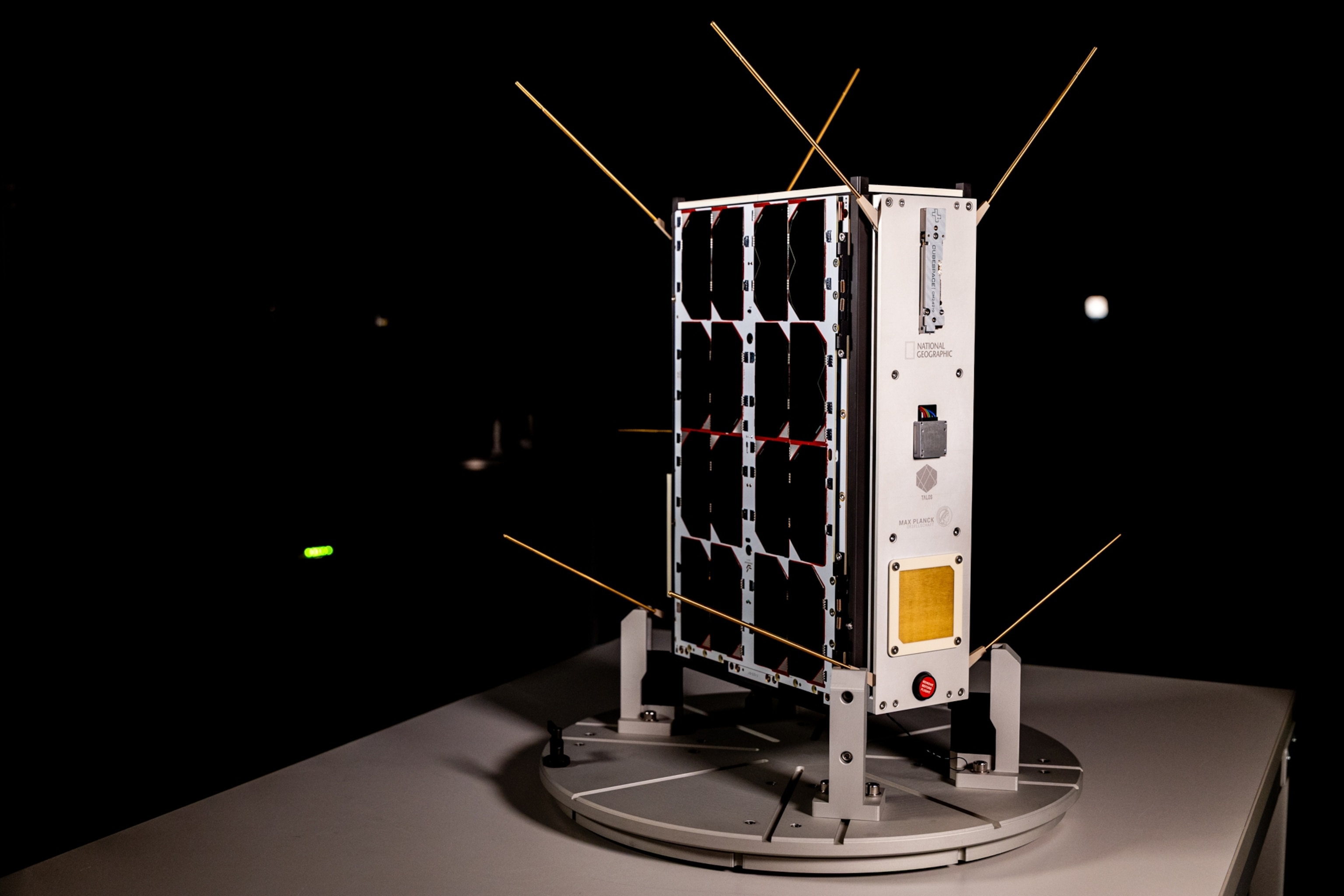 A cube-shaped nanosatellite with antennas, covered in dark solar panels, sits on a circular platform against a dark background