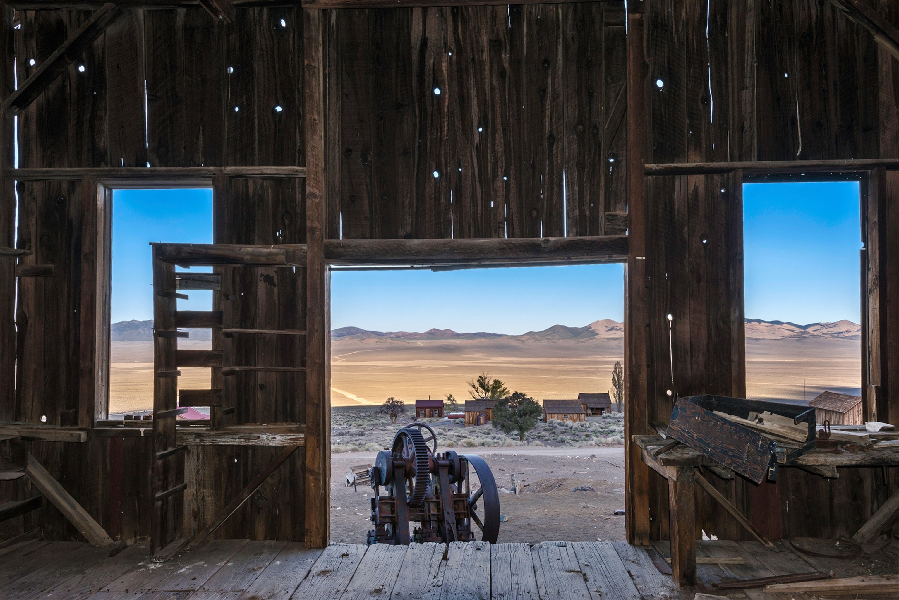 A view from inside an old abandoned wooden building looking out on a desert landscape with mountains in the distance. Rusted equipment visible outside the doorway and across the land.