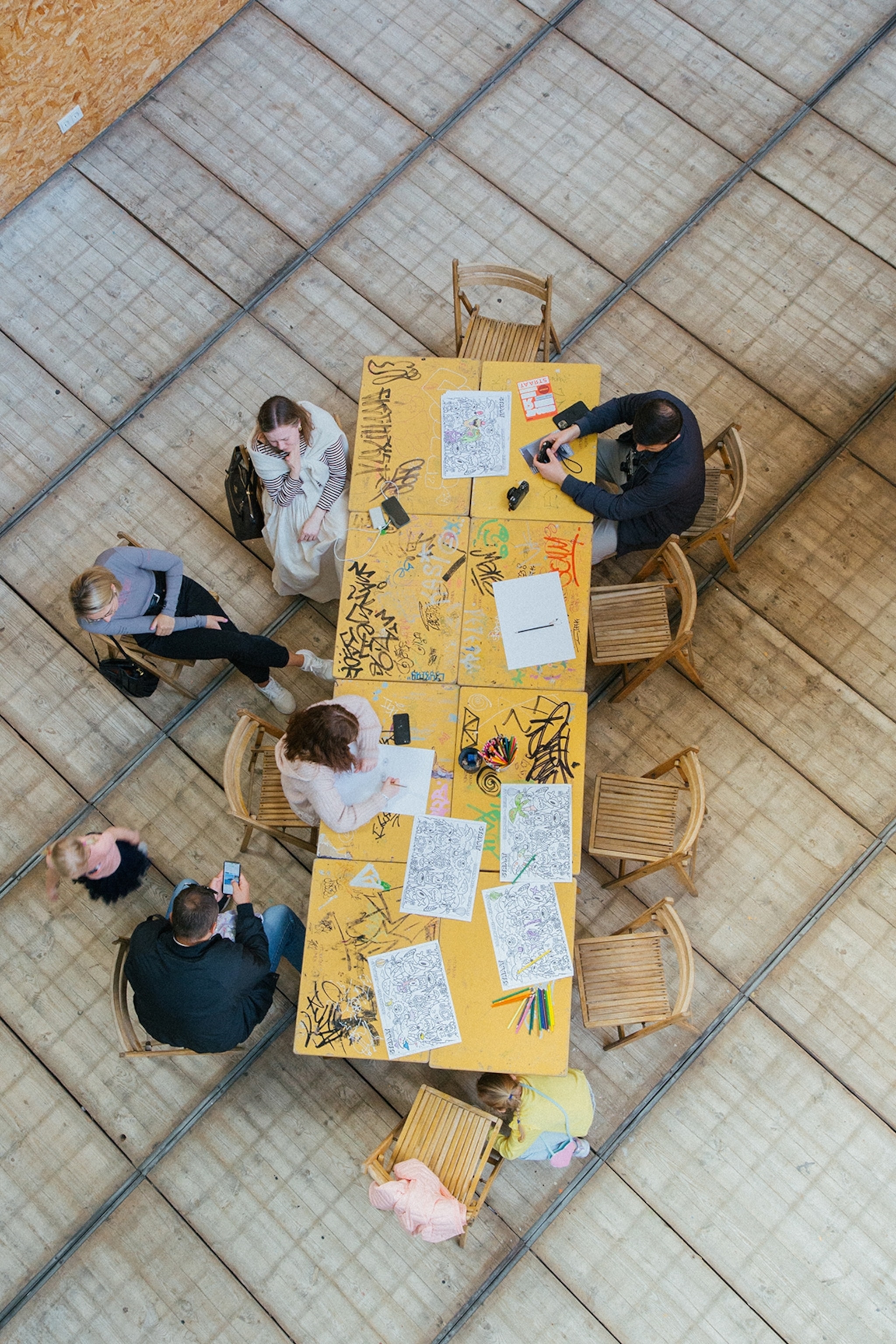 A bird's eye shot of a long, graffitied table with visitors drawing their own designs on a printed paper.