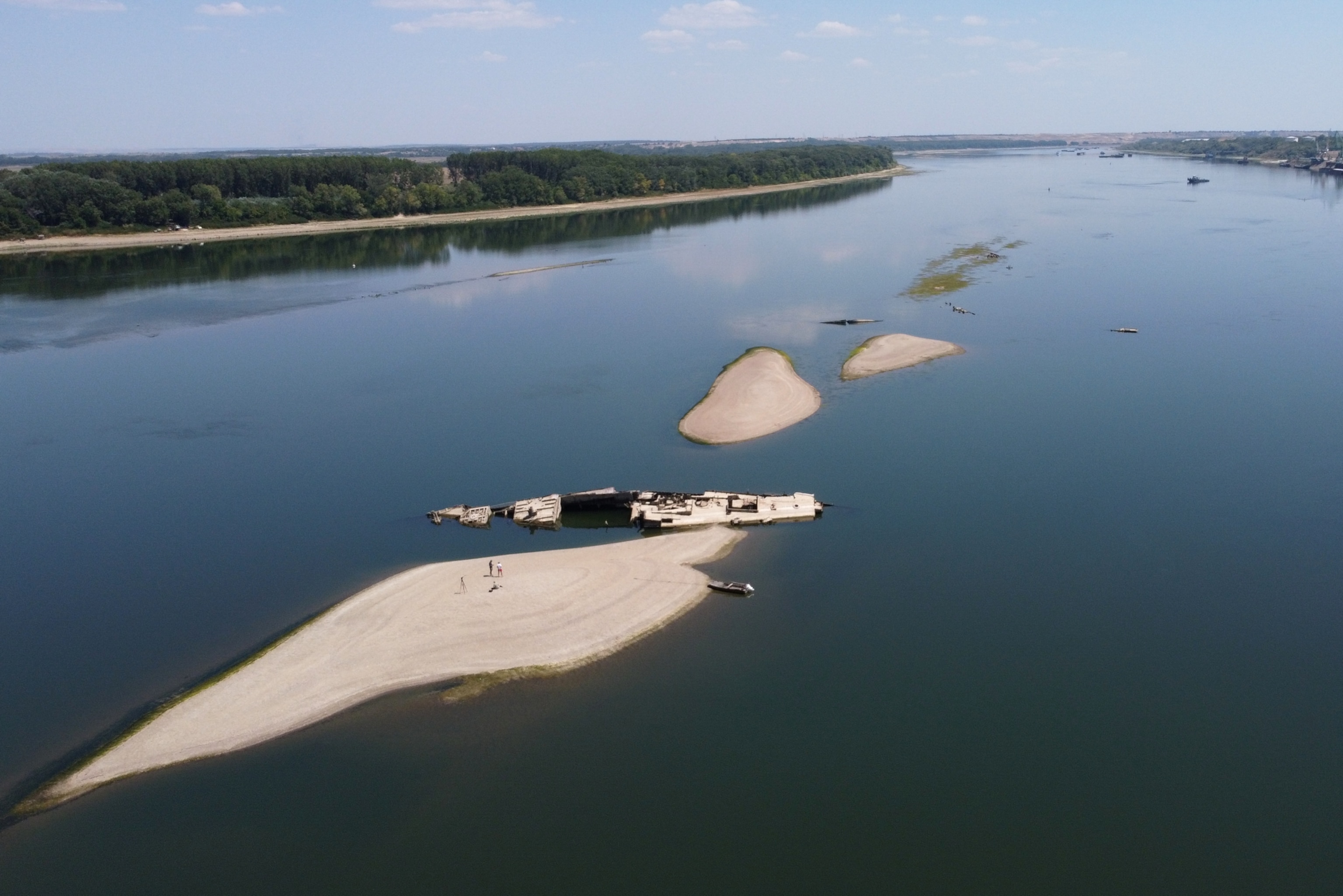 An aerial view of a wide river with exposed sandbars and the rusted, partially submerged wreckage of a large ship resting near a sandy island shore.
