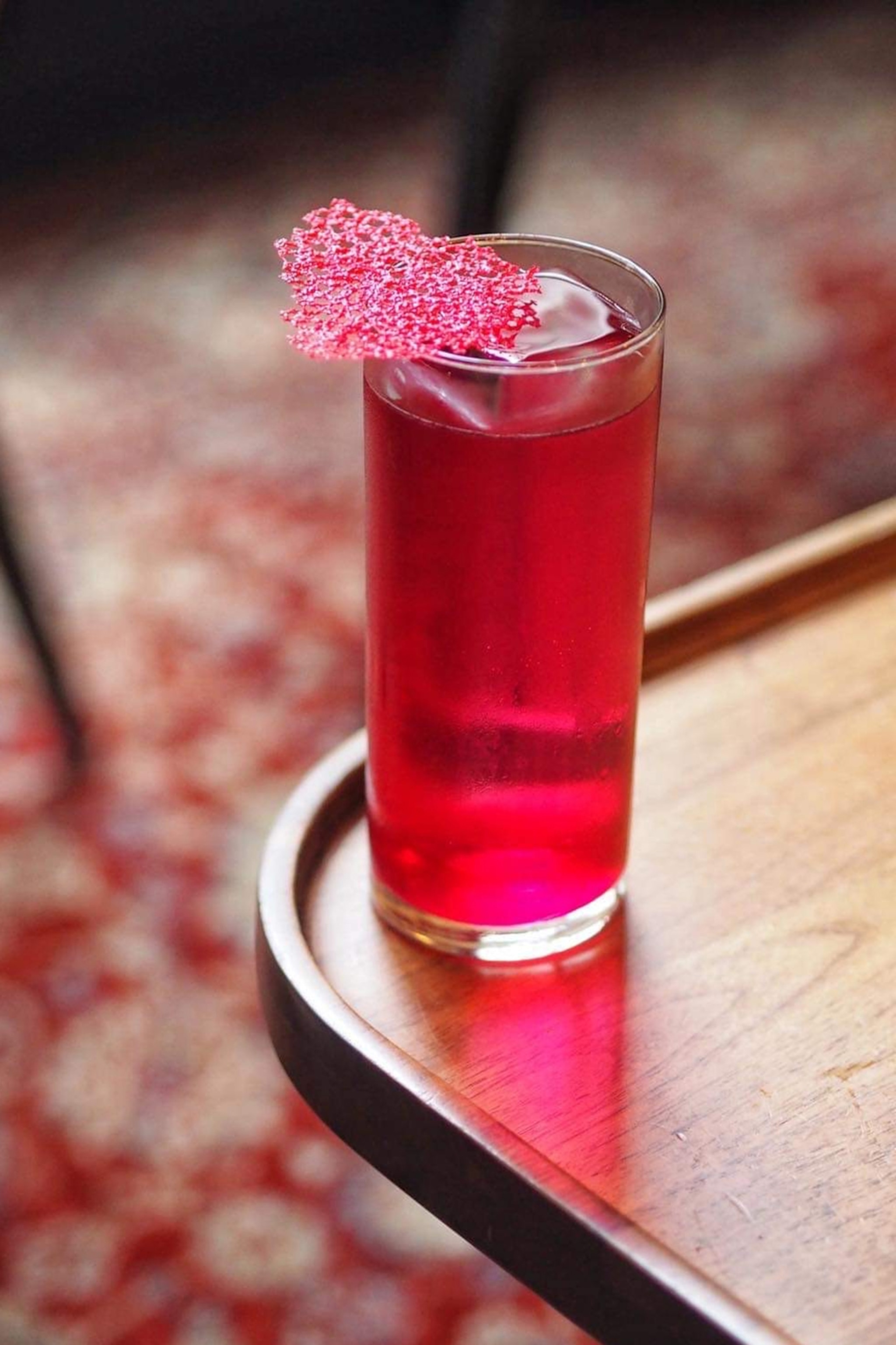 A close-up shot of a bright pink yet clear cocktail in a tall glass on the edge of a rounded, wooden bar table.