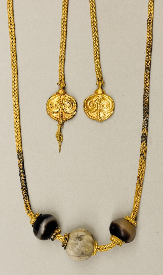 Ένα υφαντό χρυσό κολιέ αντικατοπτρίζει στυλ που συναντώνται στην Κρήτη και στην περιοχή της Πελοποννήσου, την καρδιά του μυκηναϊκού πολιτισμού. Είναι στολισμένο με χάντρες, δύο από αχάτη και μία από φαγεντιανή, και δύο χρυσά τελάρα.