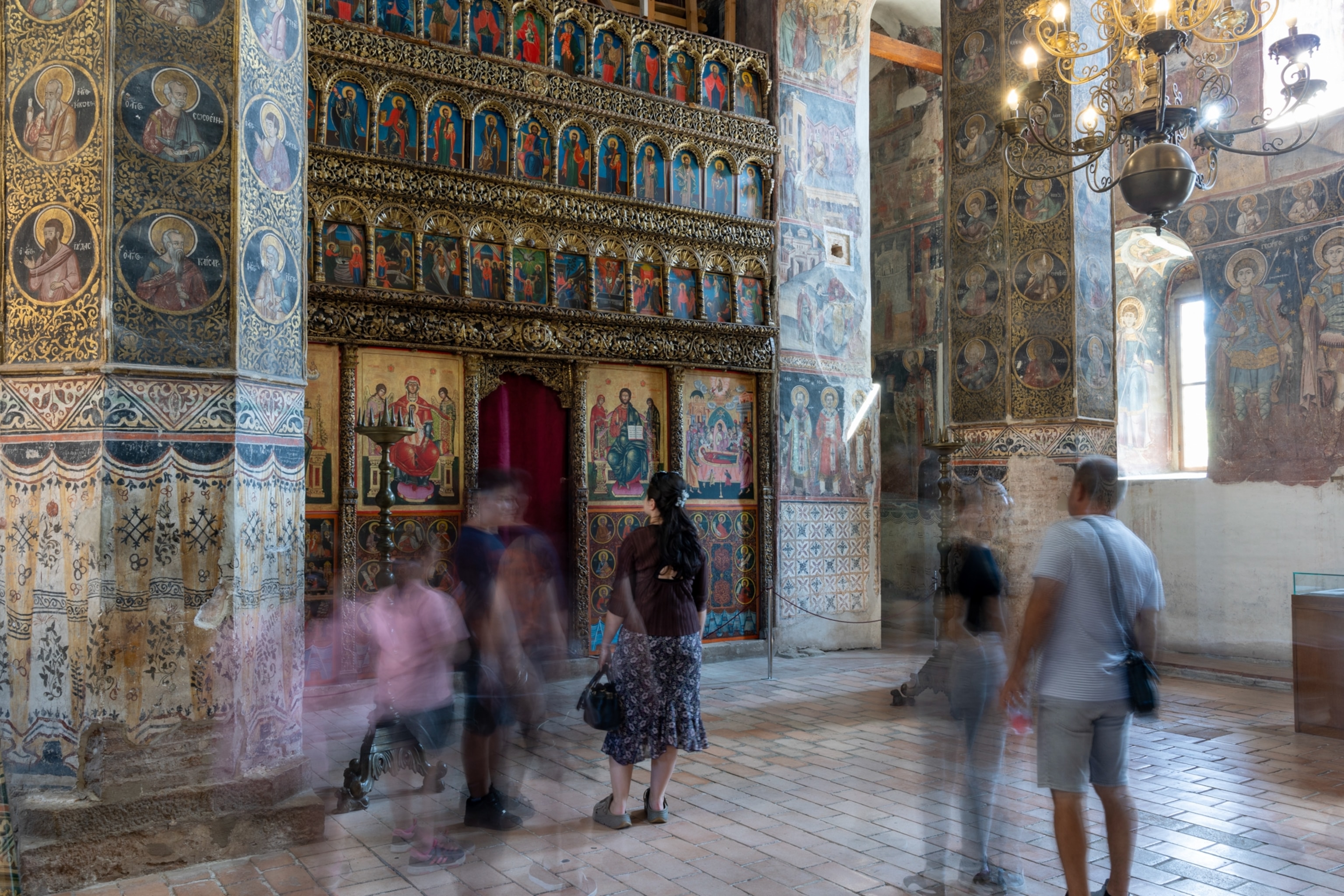 Interior of the Biserica Domnească (Princely Church) in Curtea de Argeș, with walls covered in painted biblical icons and blurred images of people in front.
