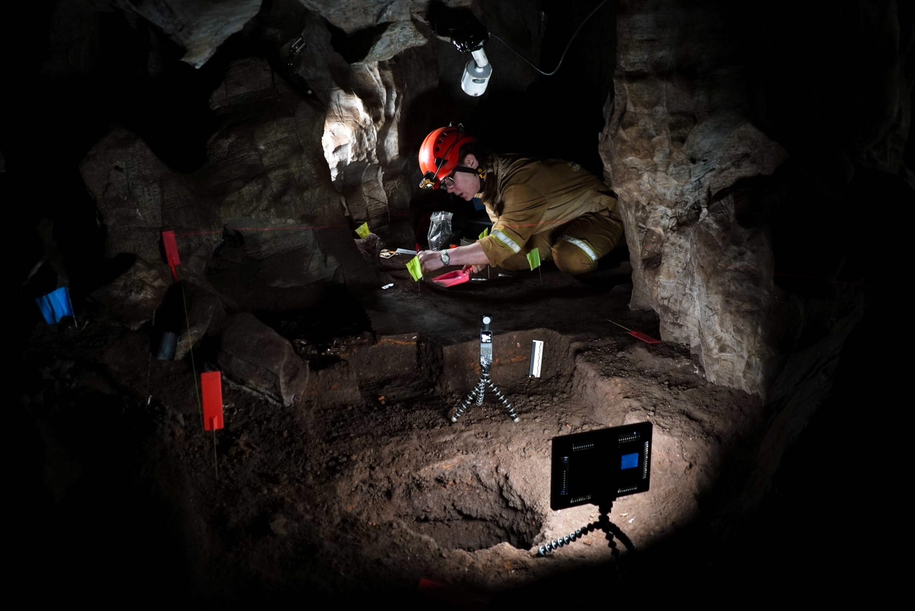 Archaeologist wearing a helmet and gloves examines artifacts in a dimly lit cave. The excavation site is surrounded by rocky walls and spotlights.