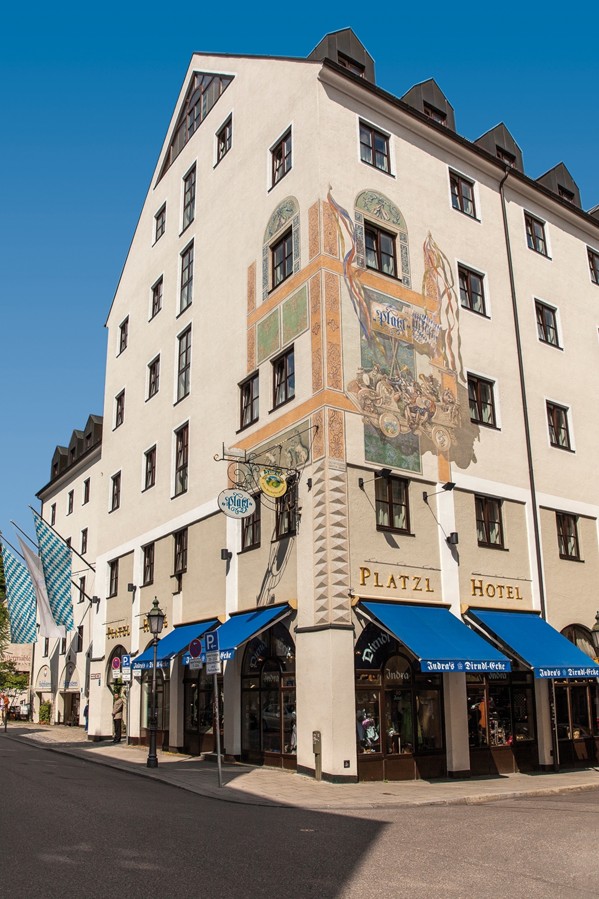 The exteriors of an unassuming building in Munich, with a medieval mural painted onto one corner of the building and sunshades rolled out over the entrance to a hotel.
