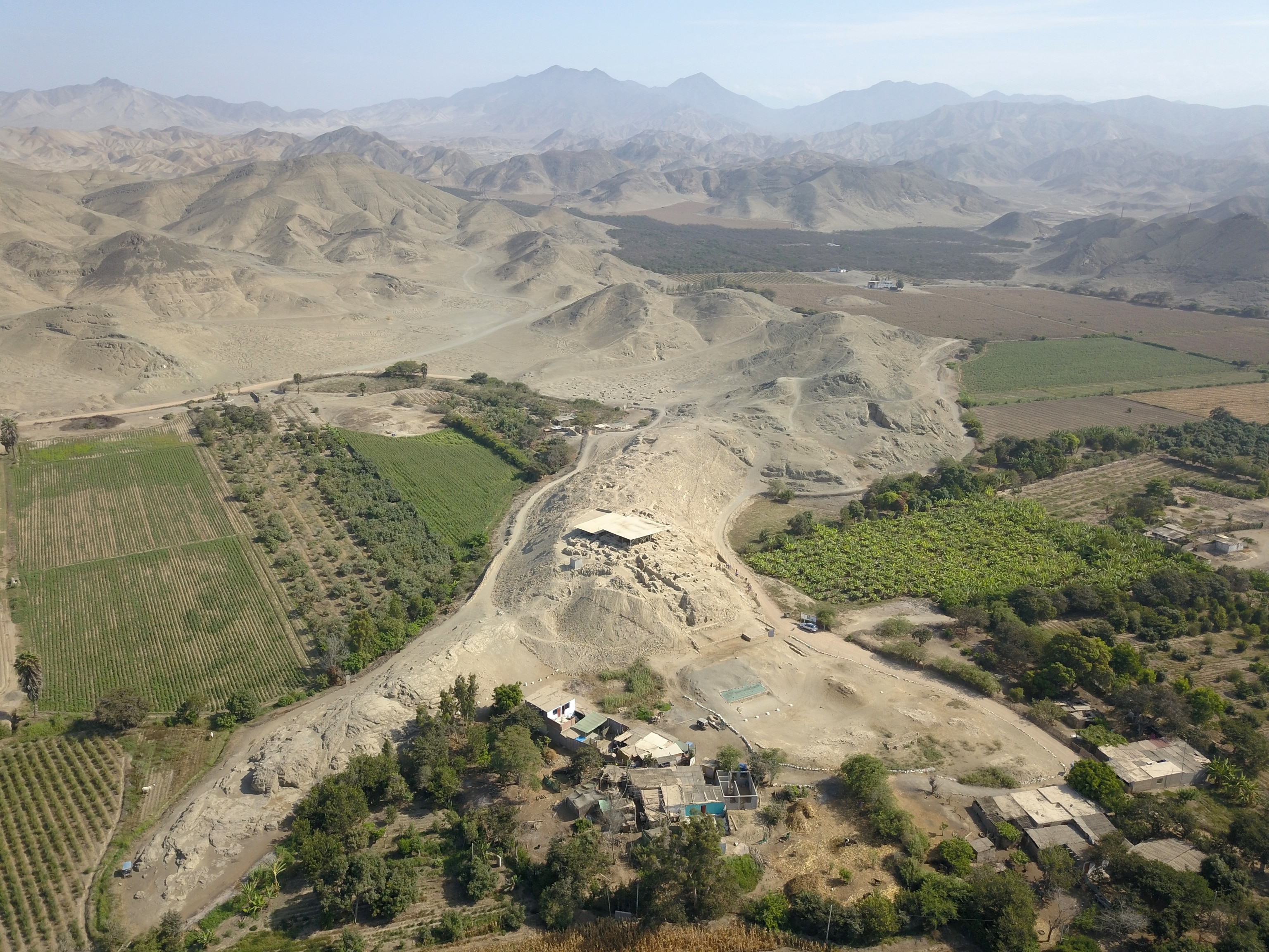 An aerial view of hilly landscape with rugged mountains in the background and a cluster of buildings nestled between verdant fields.
