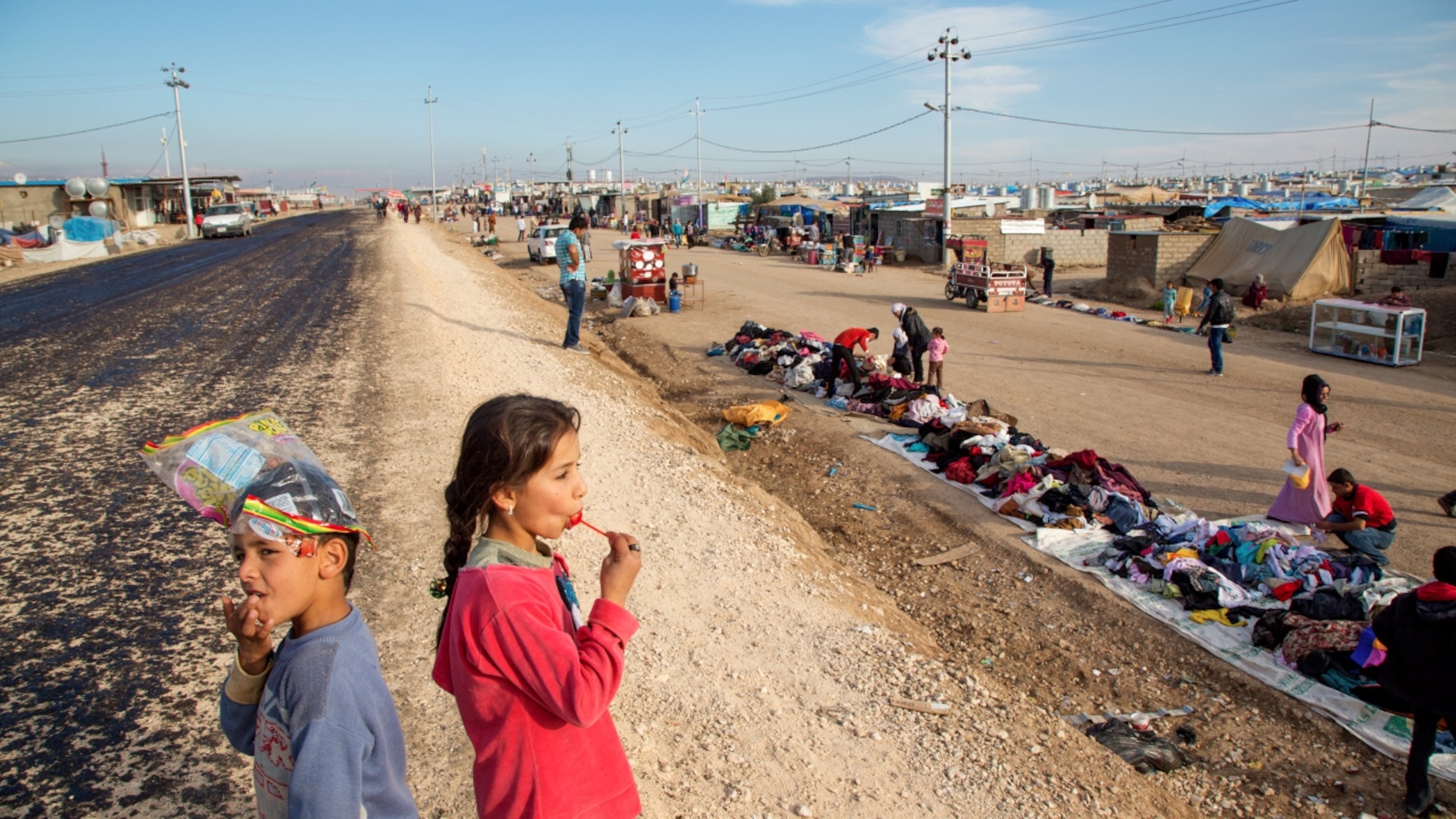A young girl enjoys a lollipop while watching shoppers in the Domiz Camp for Syrian Refugees just outside of Dohuk, Iraq.
