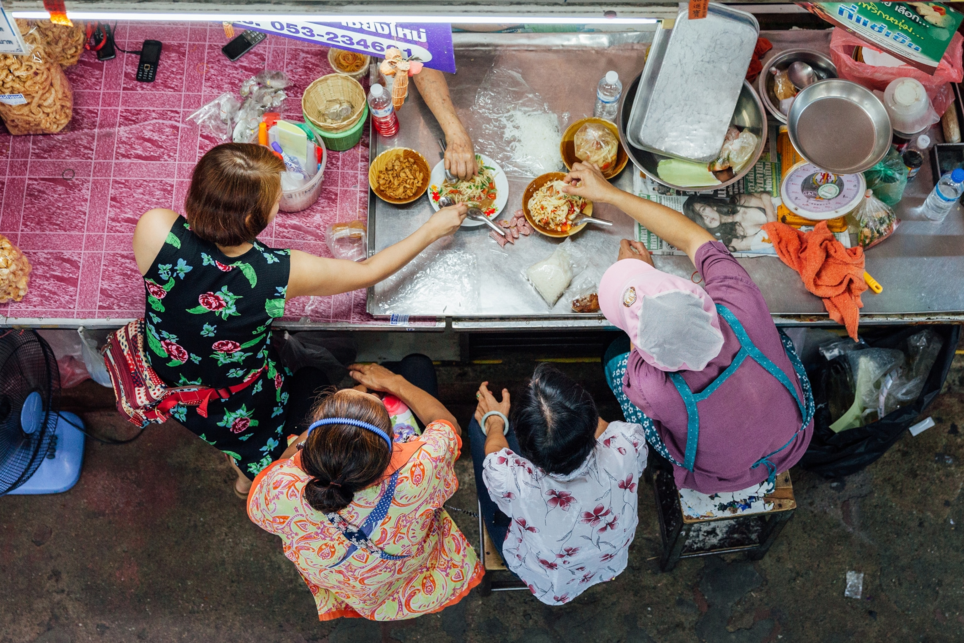 A birds-eye perspective on four Thai women sharing bowls of food in the kitchen of a street food stall.