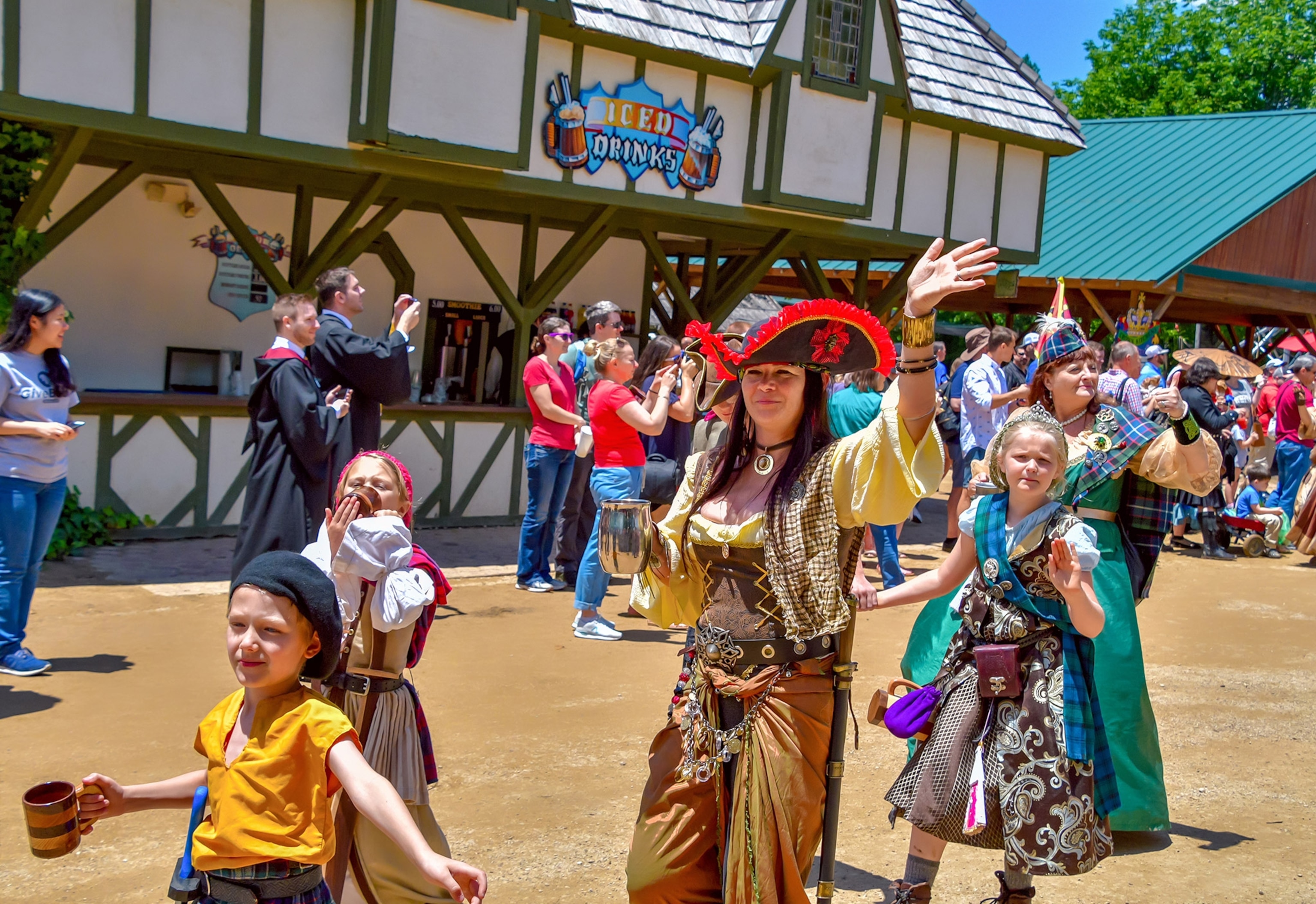A group of people in varied Medieval costumes walks and stands around a dirt road in bright sunshine