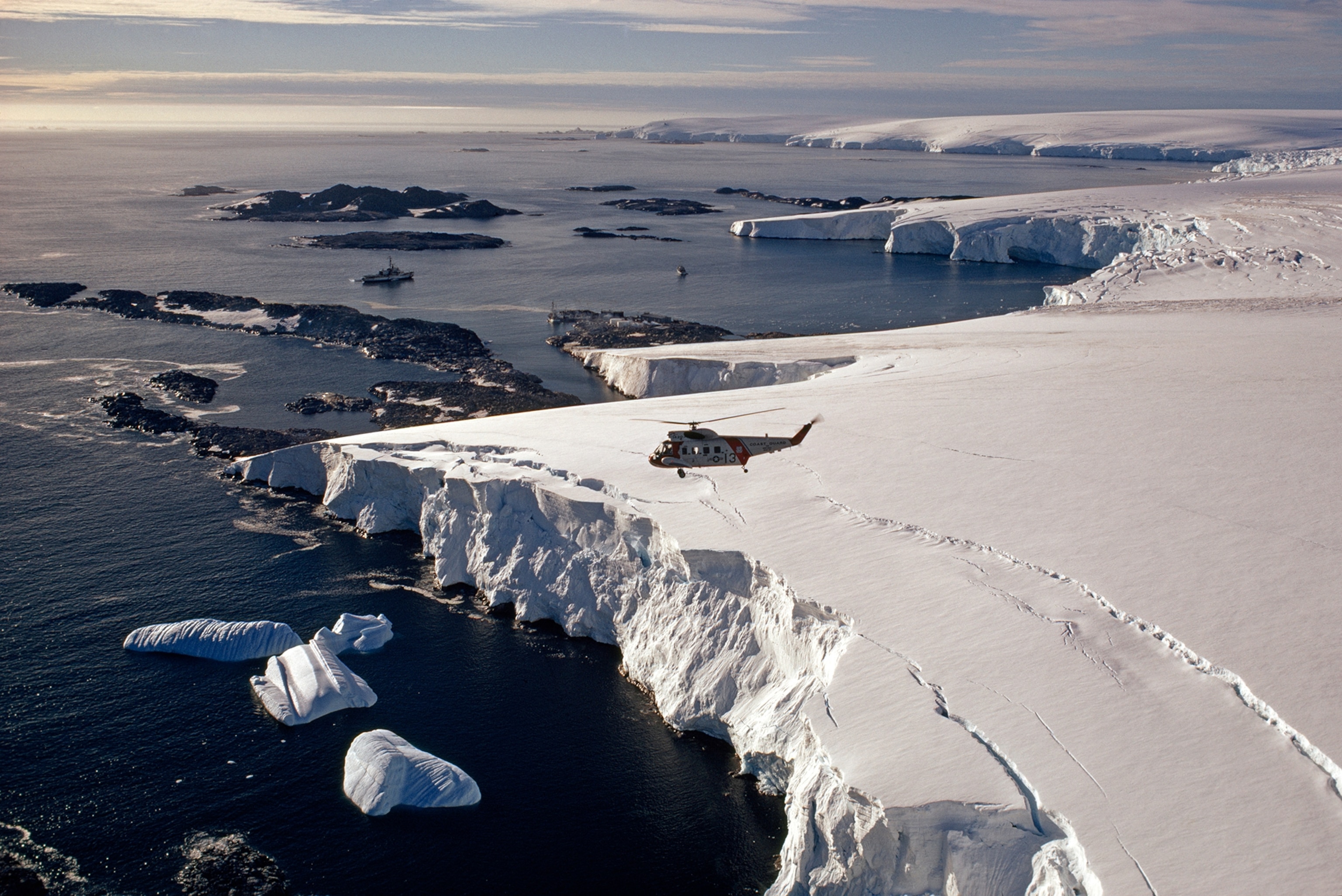Between snows and seas, inconspicuous Palmer Station clings to a tiny icefree spit on Anvers Island, at center. Icebergs strew the water, and crevasses promise more. The icebreaker Westwind and a smaller research ship, Alpha Helix, anchor offshore; Hero lies at the dock. A Westwind helicopter surveys the scene. One of Antarctica's four year-round U. S. stations, Palmer is the closest to South America. Built at water's edge, it seems exposed, but summer storms are few, and winter congeals Arthur Harbor into an icy, rigid plain.