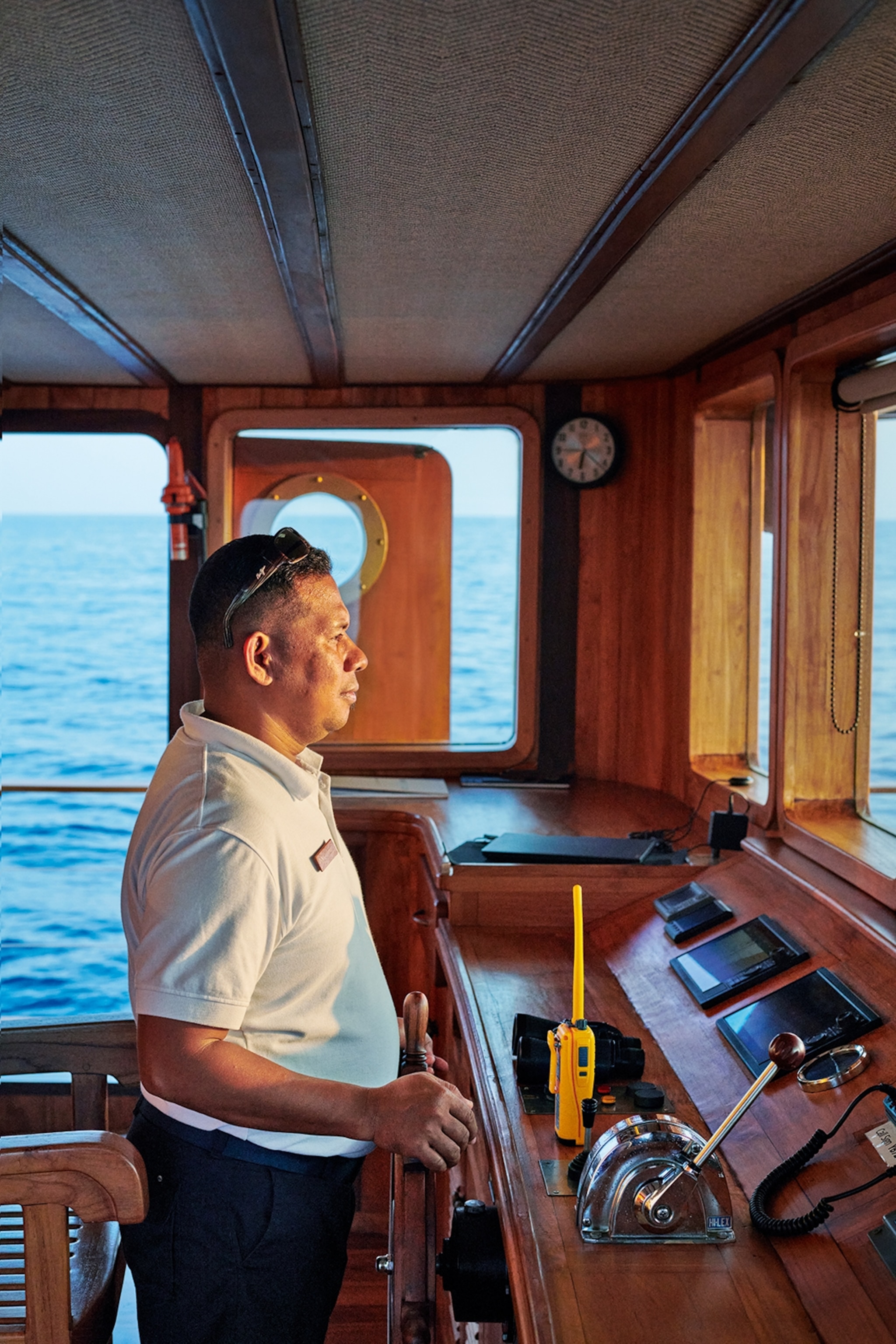 A side profile of a local, Indonesian male captain staring ahead from his wooden steering wheel towards the setting sun.