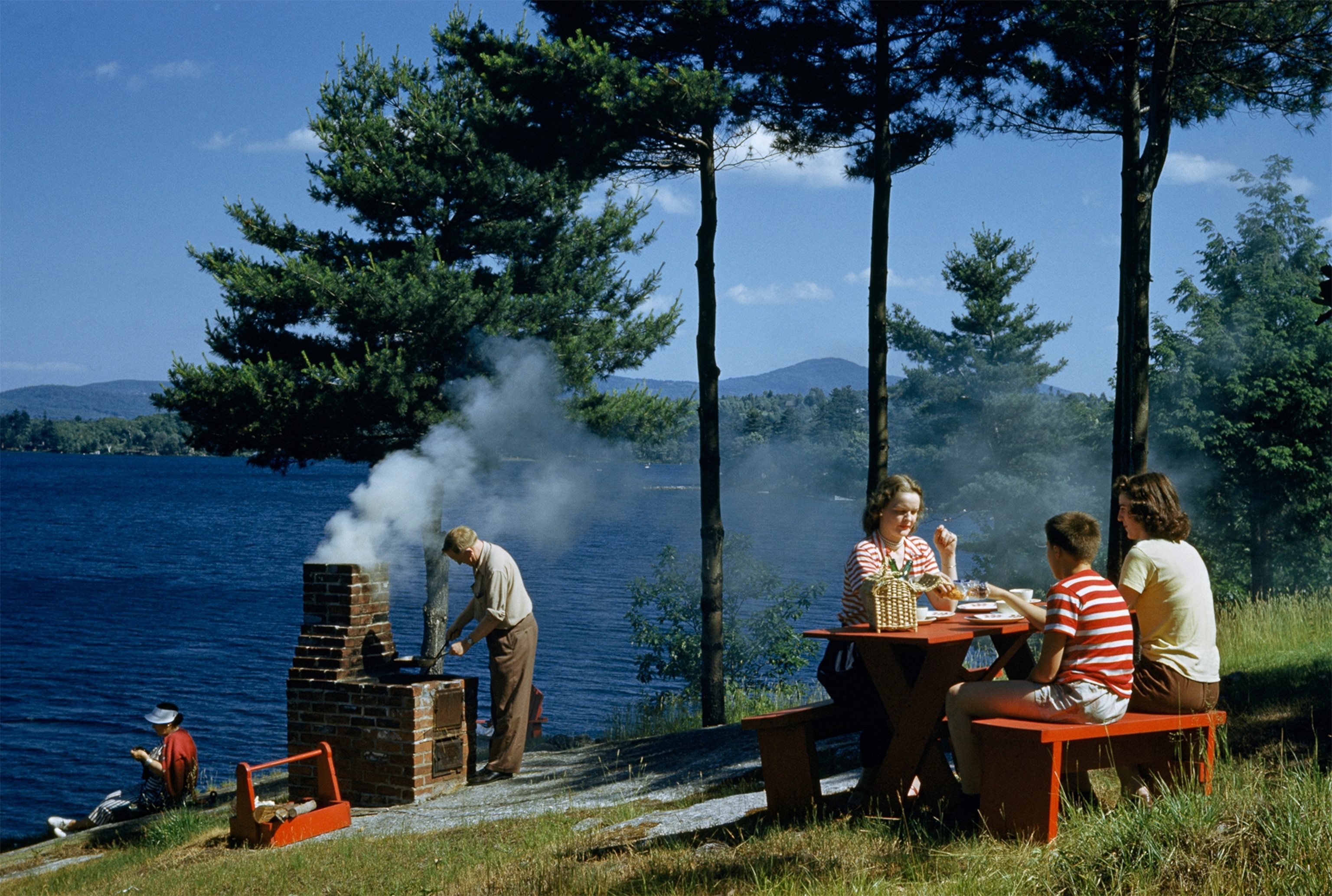 people grilling food and picnic on hillside overlooking a lake in 1948