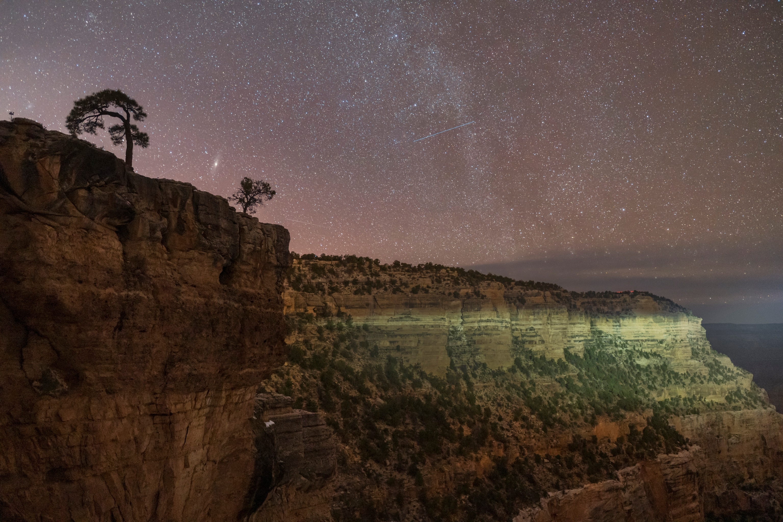 A side of the Grand Canyon is illuminated by a passing cars headlight, with the night sky behind it full of stars