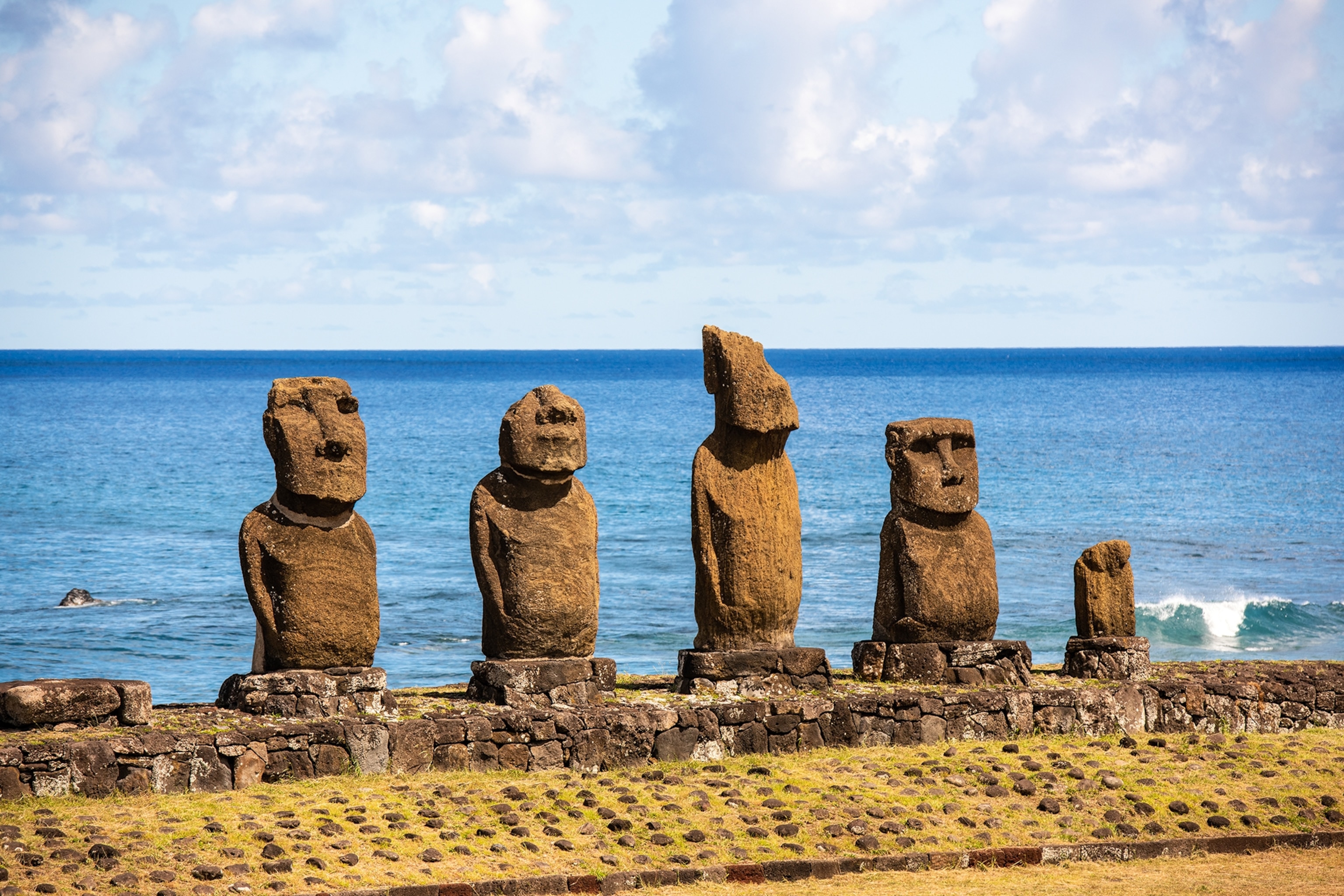 A line-up of human-like stone statues on the edge of an island with the ocean in the background.