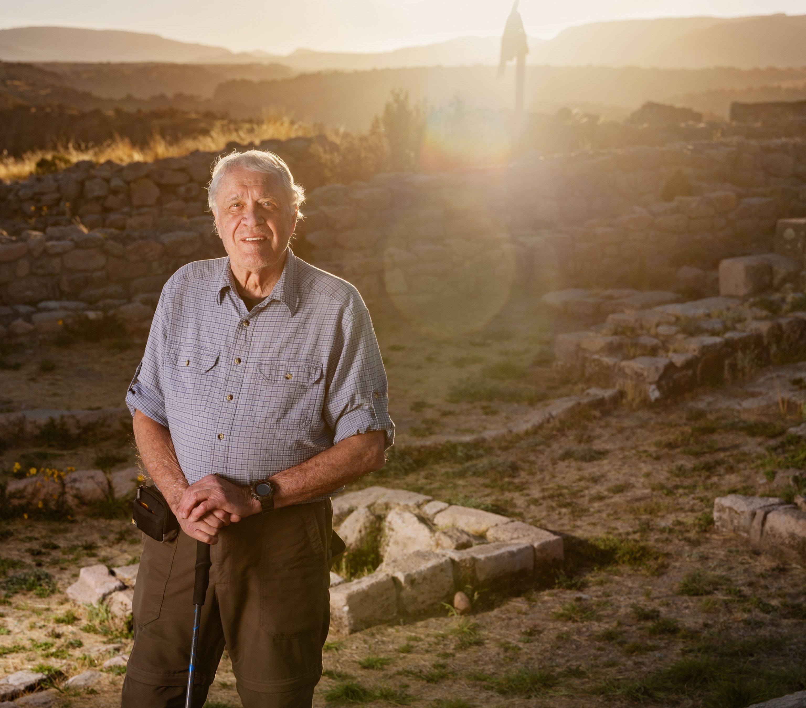 Man with his hands on walking stick stands among the stone structures at the archaeological site and looking into camera.