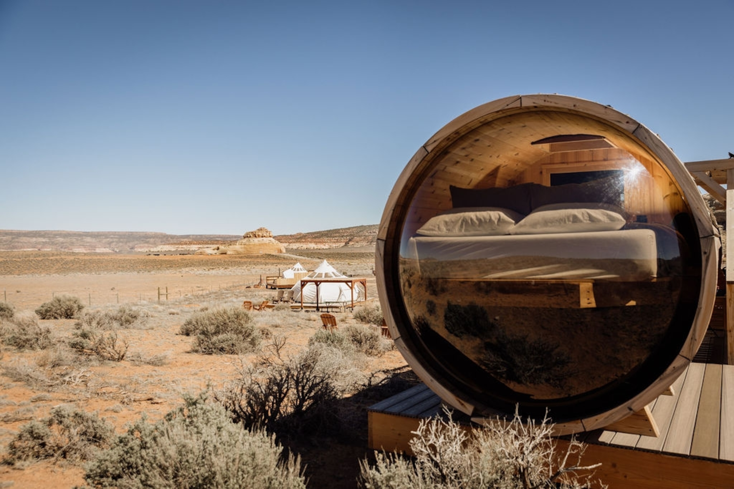 A small sleeping pod with a bed visible through fish-eye glass, sitting in a dry desert landscape with other structures in the distance