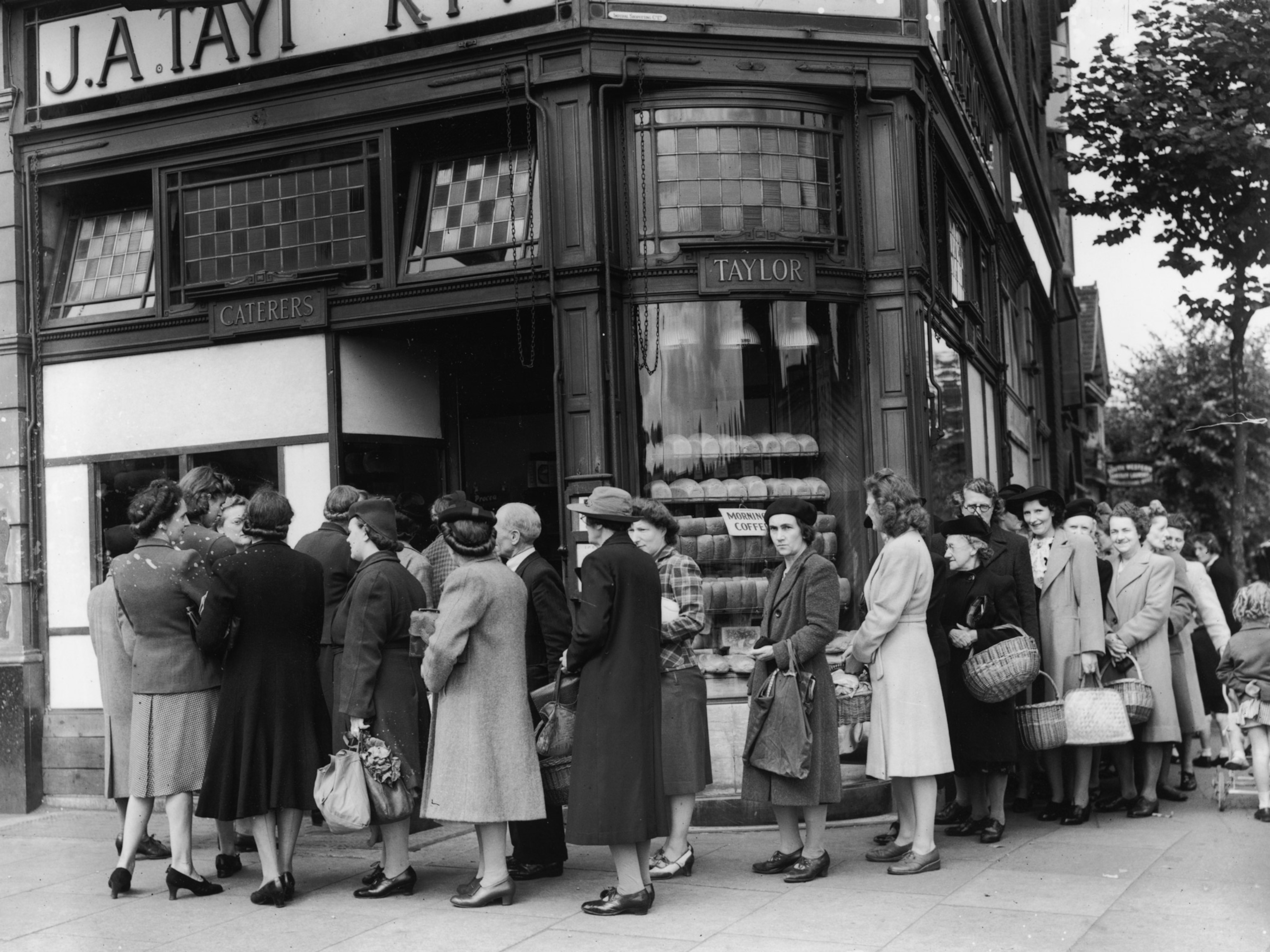 Black and white, a long queue of people wraps around a street corner, as people wait to enter a building on the left.