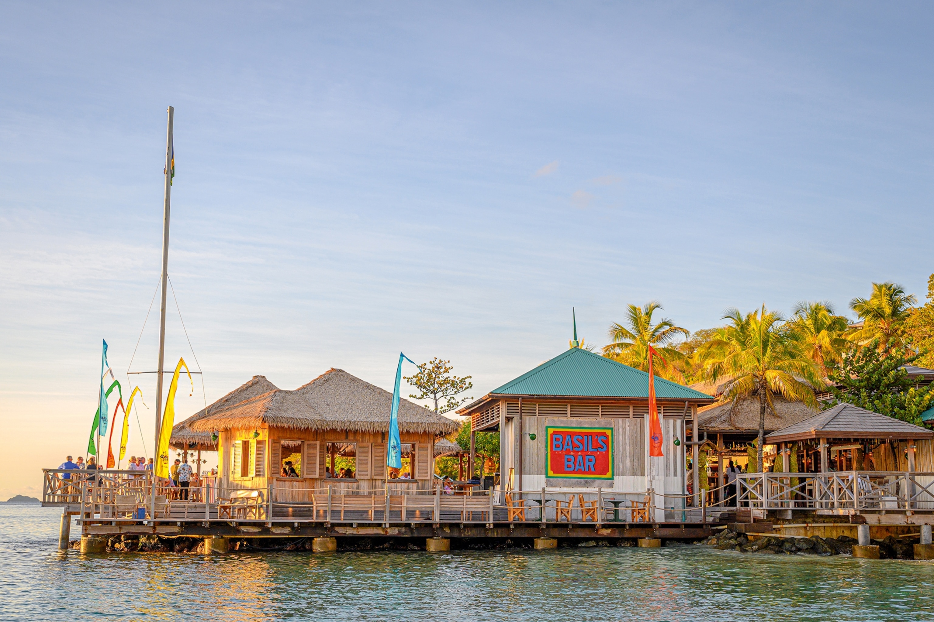 A side view onto a pier-mounted restaurant bar with straw-roofed bungalows at dusk.