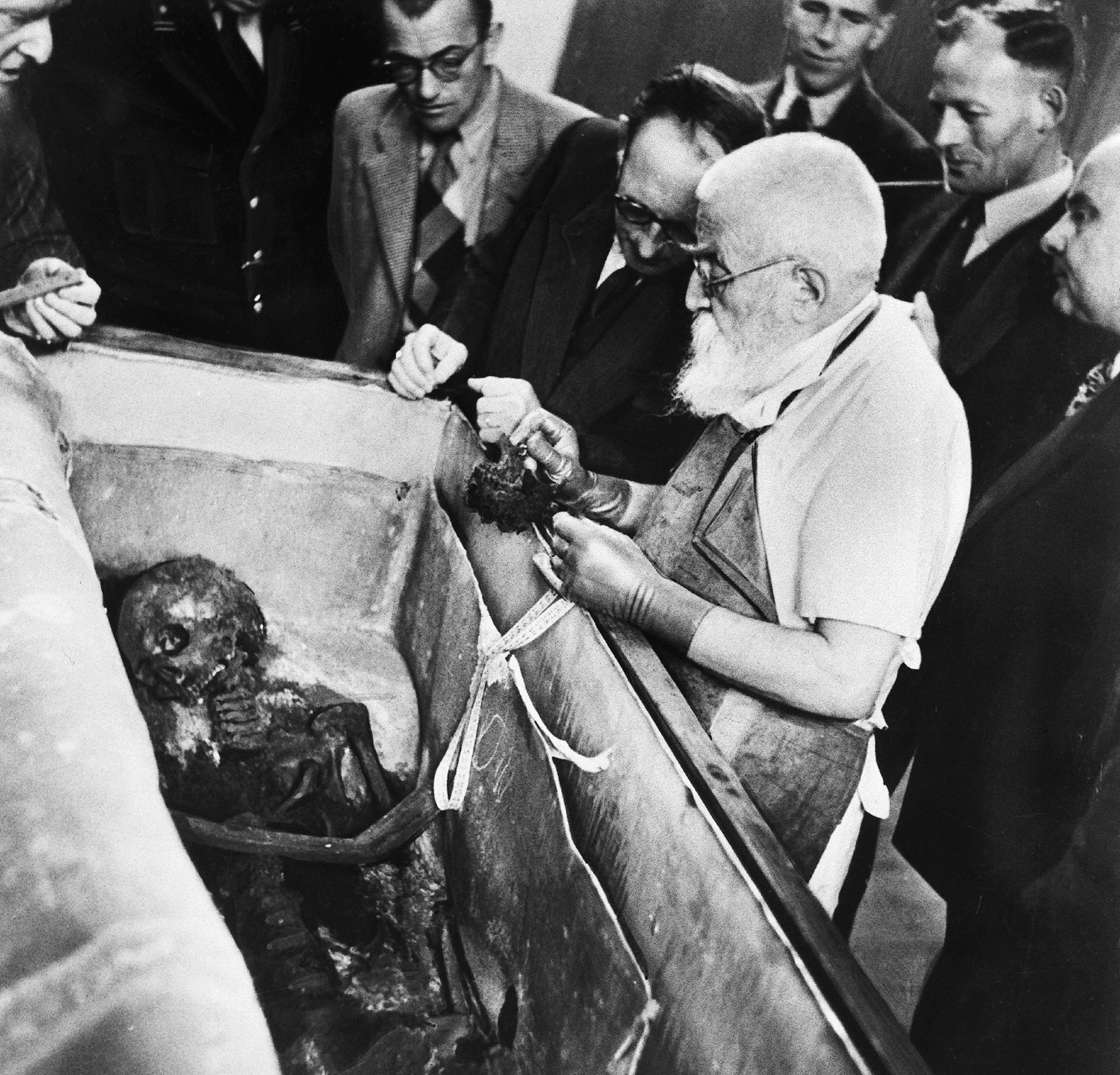 1950 black and white photo depicting a man in white shirt, apron, and gloves, standing over an open coffin with embalmed body/skeleton inside. A group of men stand around observing.