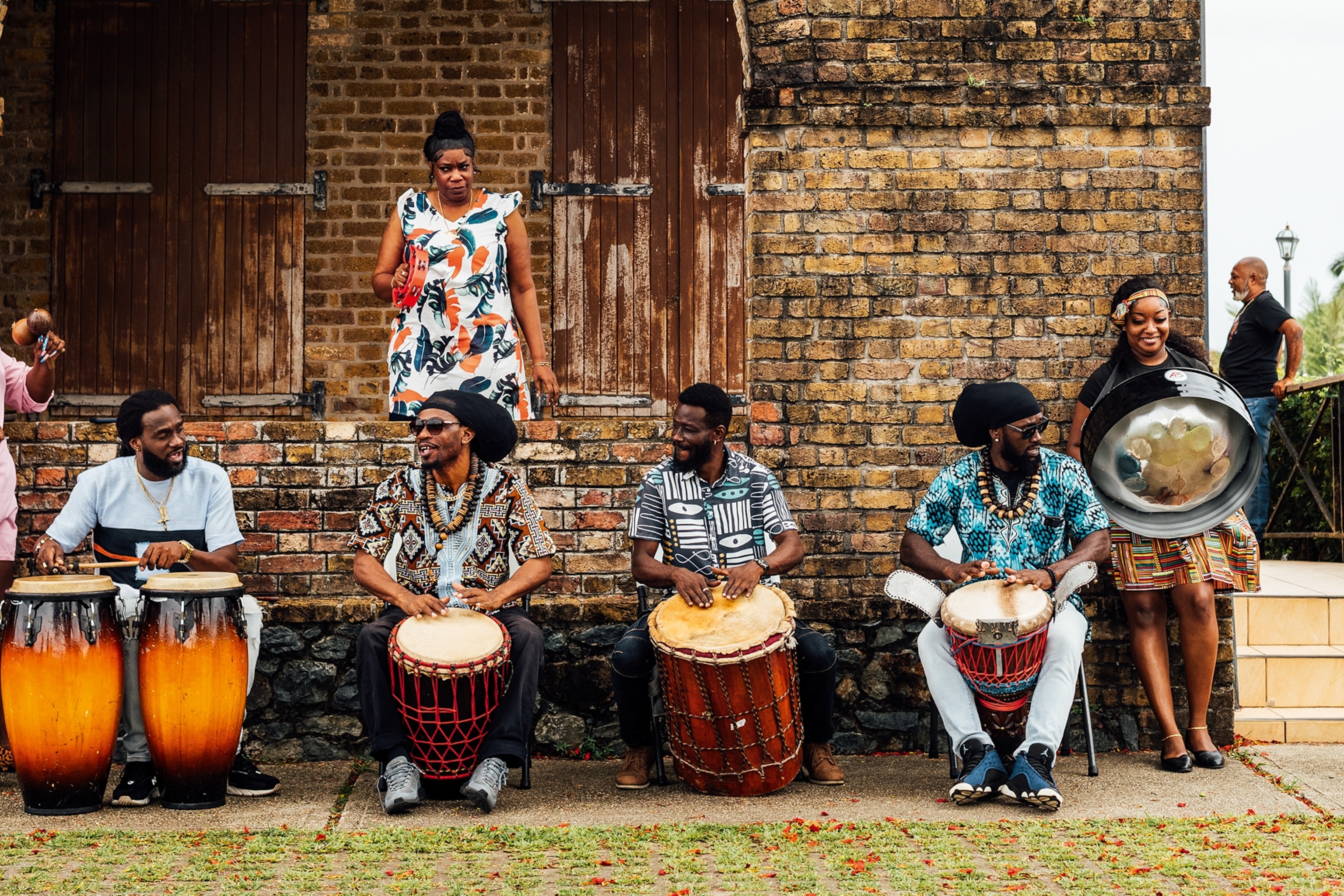 A band of four men sitting behind drums as two women join with hand-held instruments.