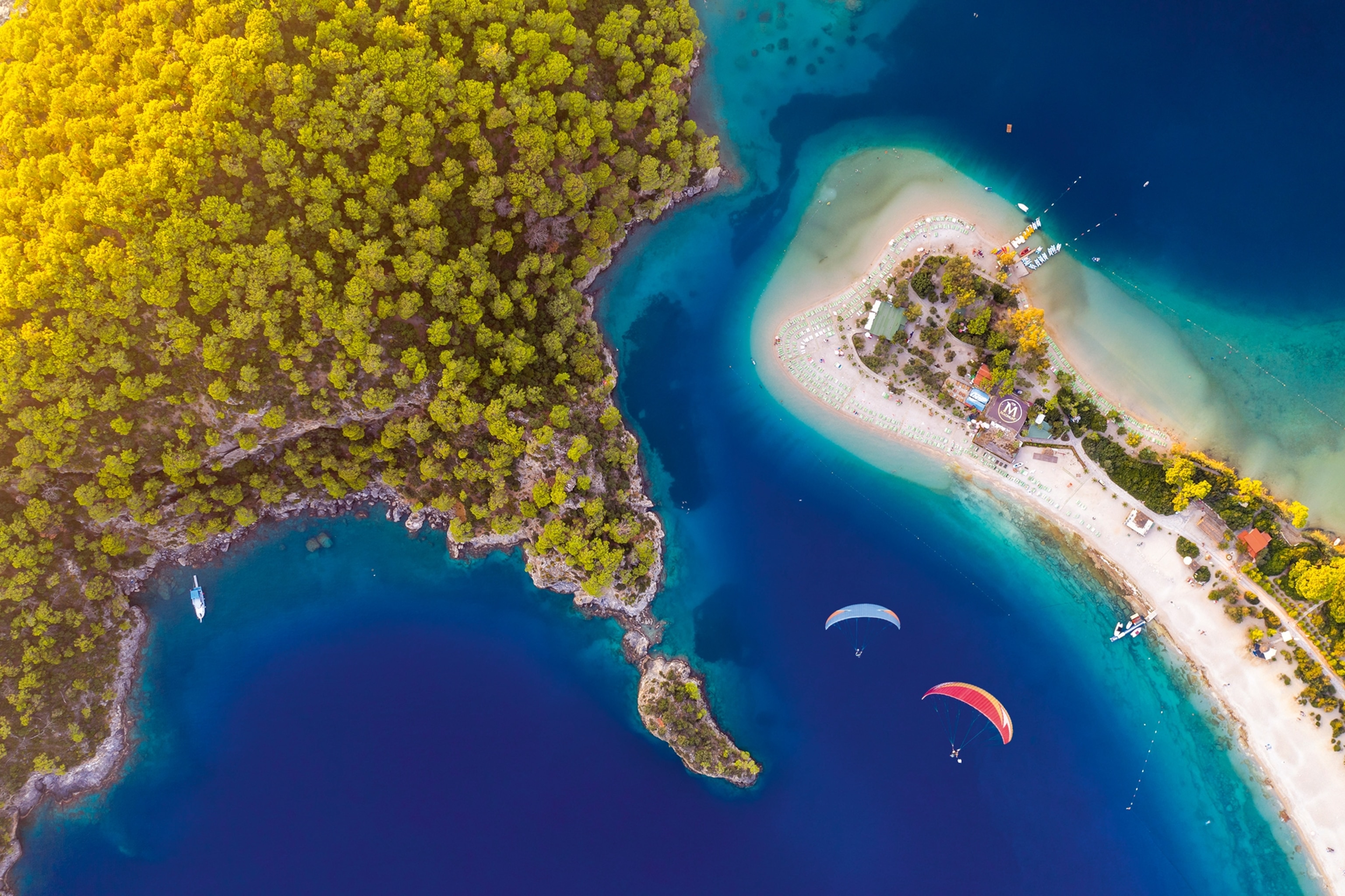 An aerial view over a beach enclave on a coast with paragliders circling above.