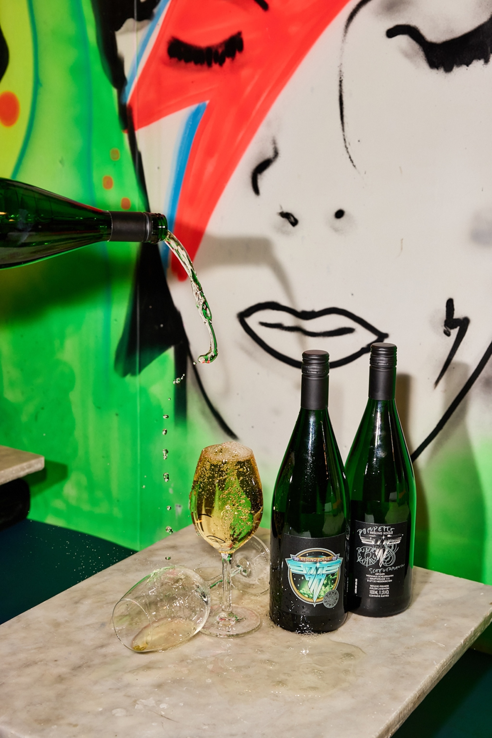 Two glasses of wine being clumsily filled with white wine to the backdrop of a David Bowie wall mural.
