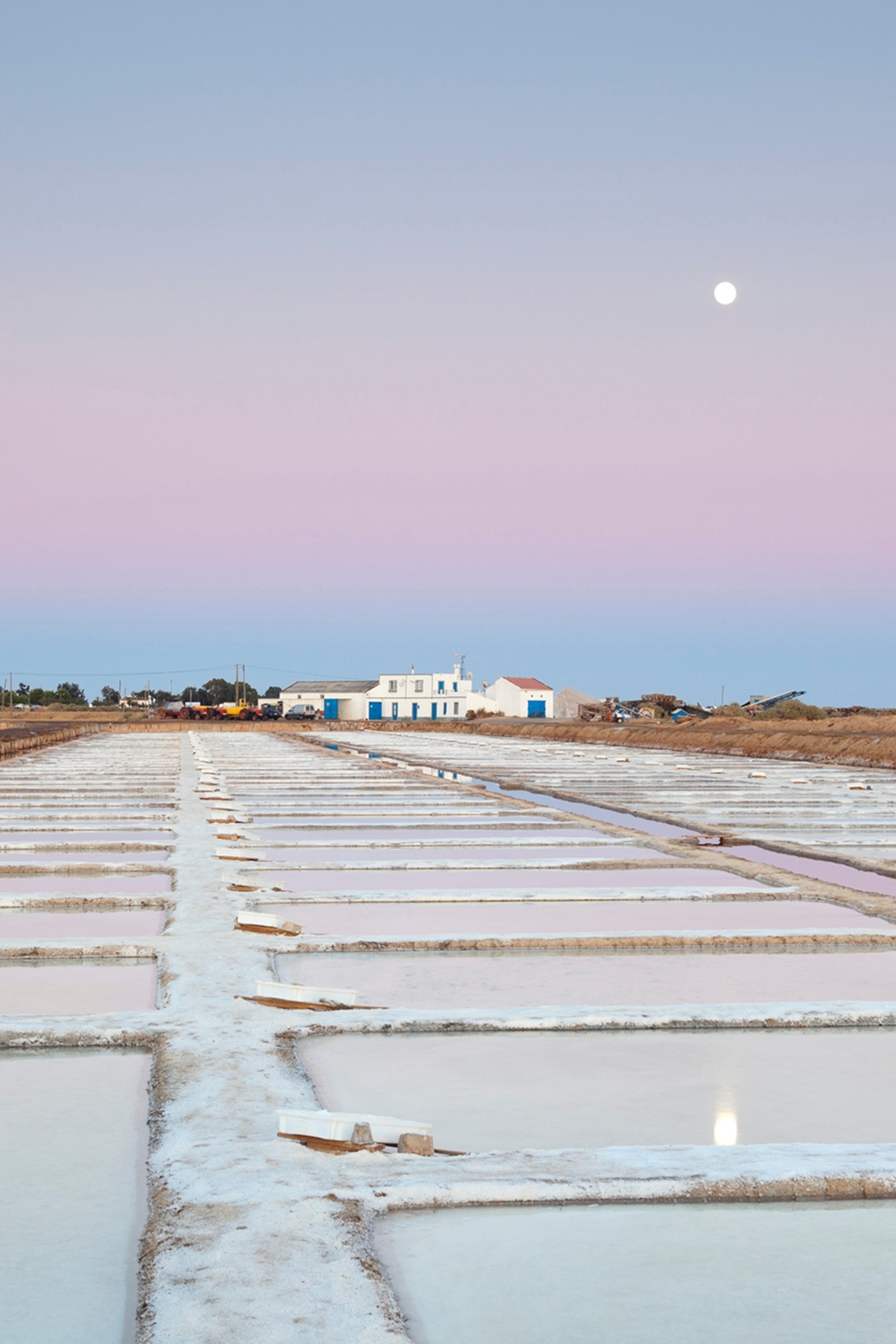 Far-stretching salt pans with a house in the distance and the moon in the sky at late dusk.