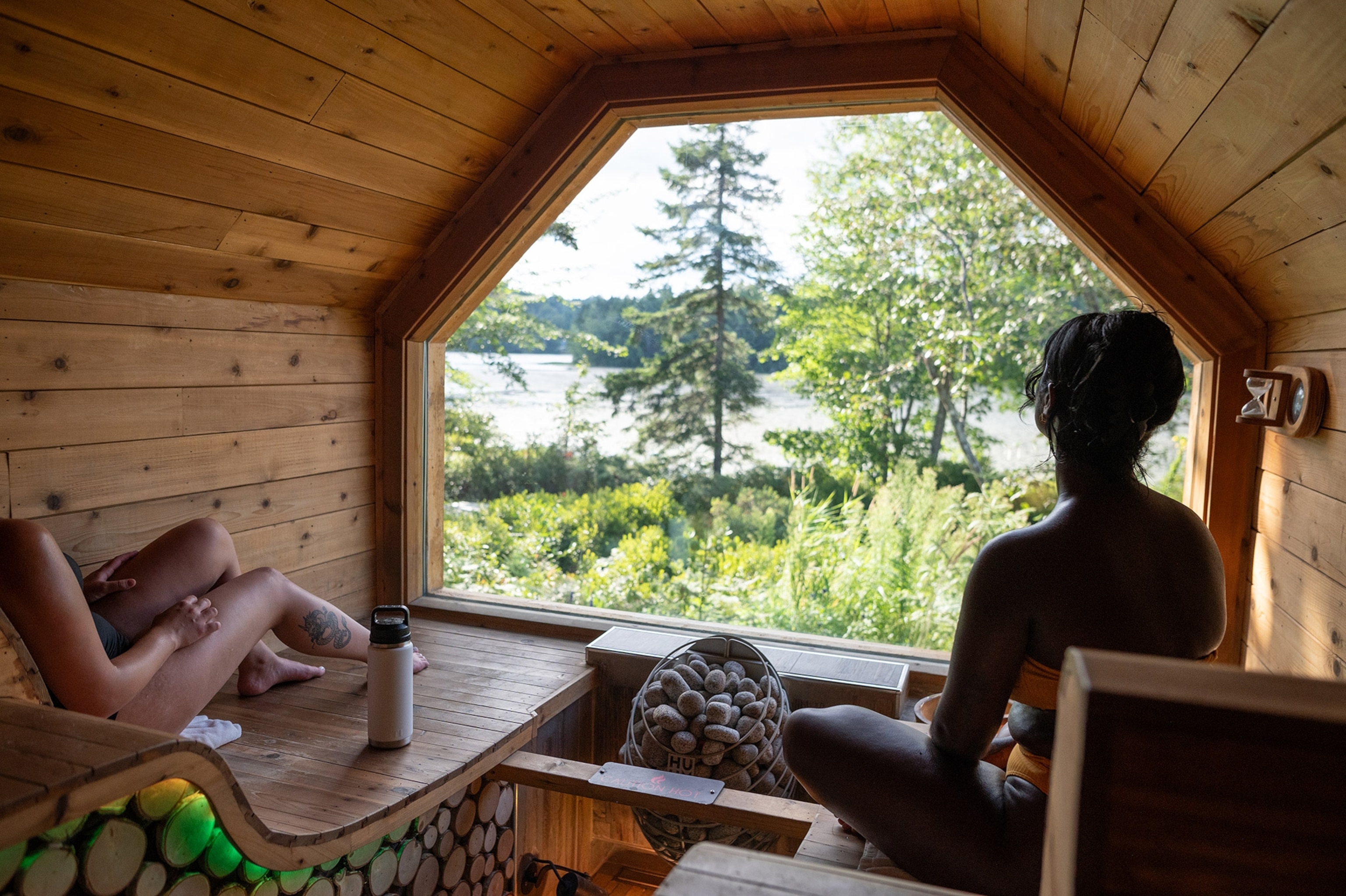 Two women sit on lounges in a small wood-paneled space, facing a large window looking out into wooded natural area