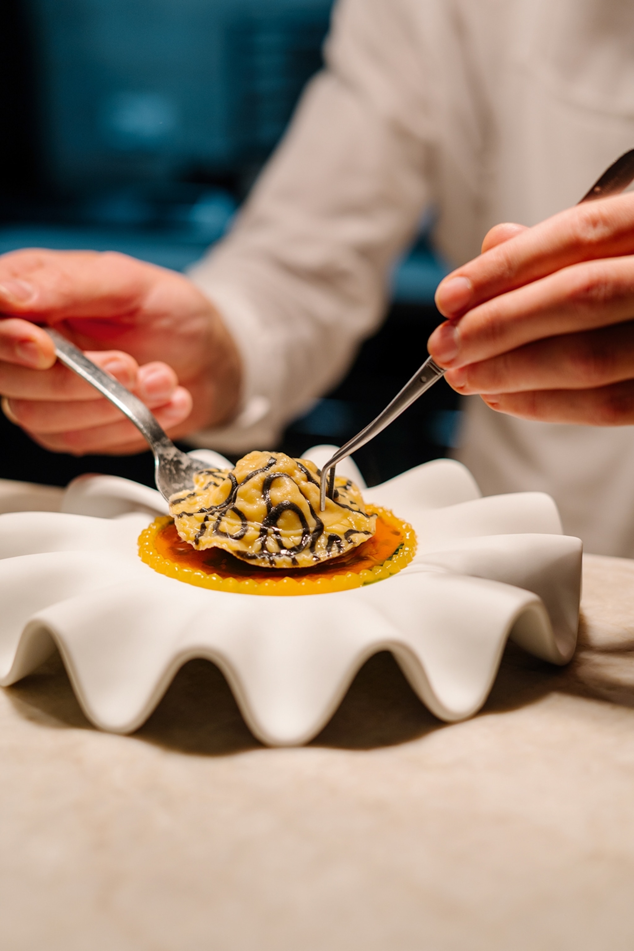 A close-up of two hands lowering a single ravioli with pliers onto a small, wavy plate in a fine-dining restaurant.