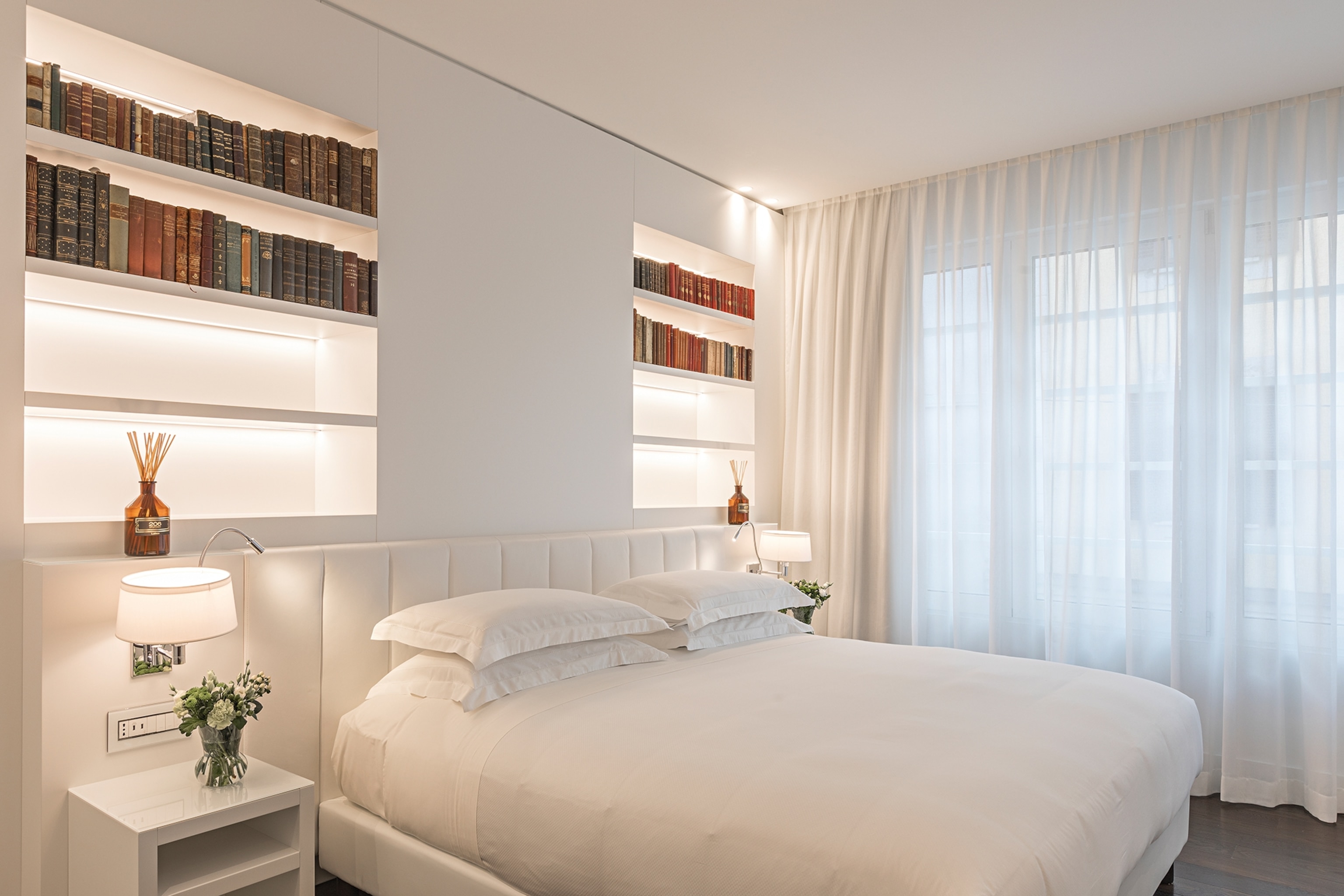 The interiors of a sleek and modern hotel bedroom with a double bed, padded wall doubling as a headboard and room perfume bottles on a shelf.