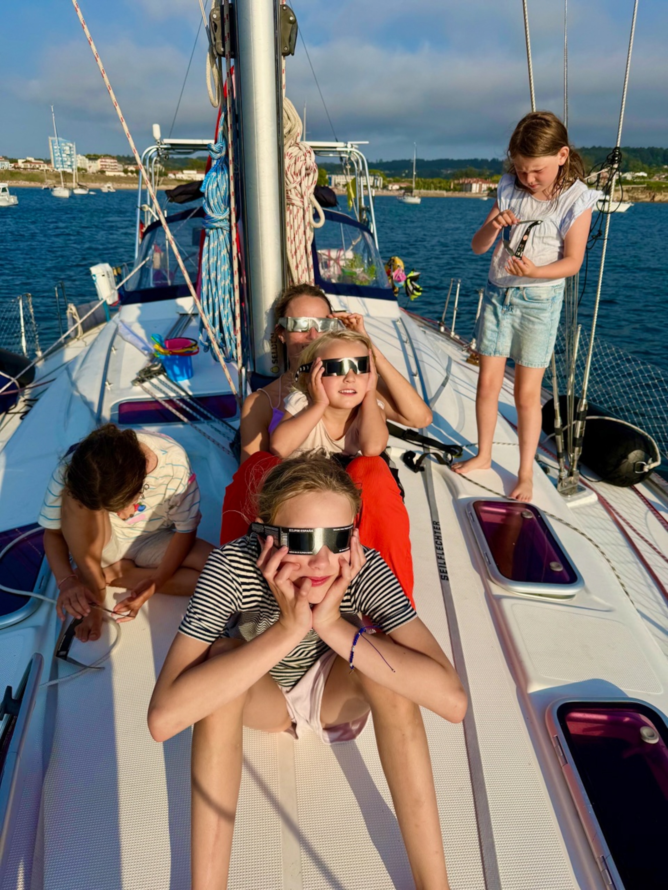 Children playfully pose in oversized, protective, eclipse-viewing glasses while on a sailboat deck, with sparkling blue water, rigging, and a sunny coastal scene behind them.
