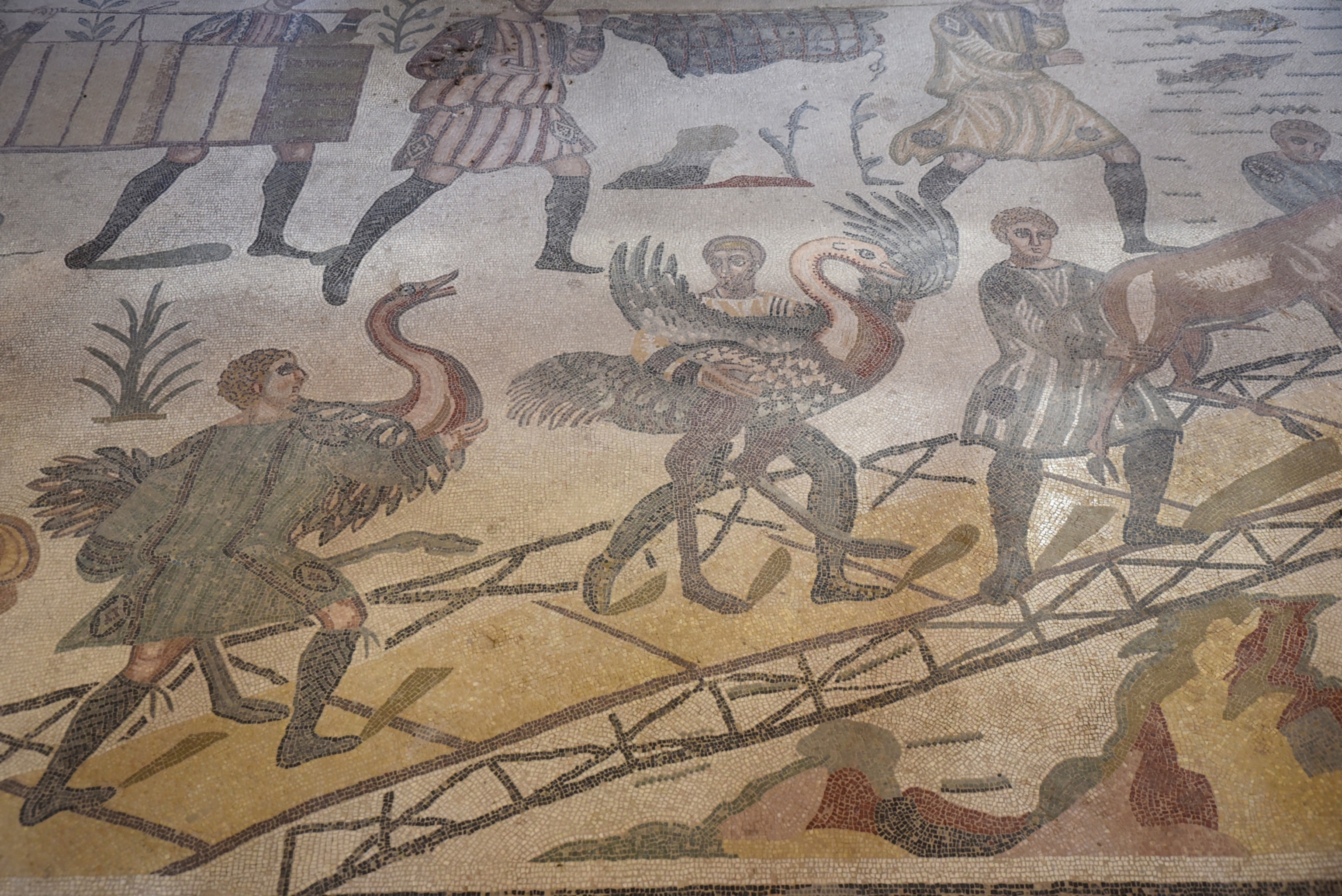 An ancient Roman mosaic shows people hunting large birds in a detailed scene made from small colored stone tiles.