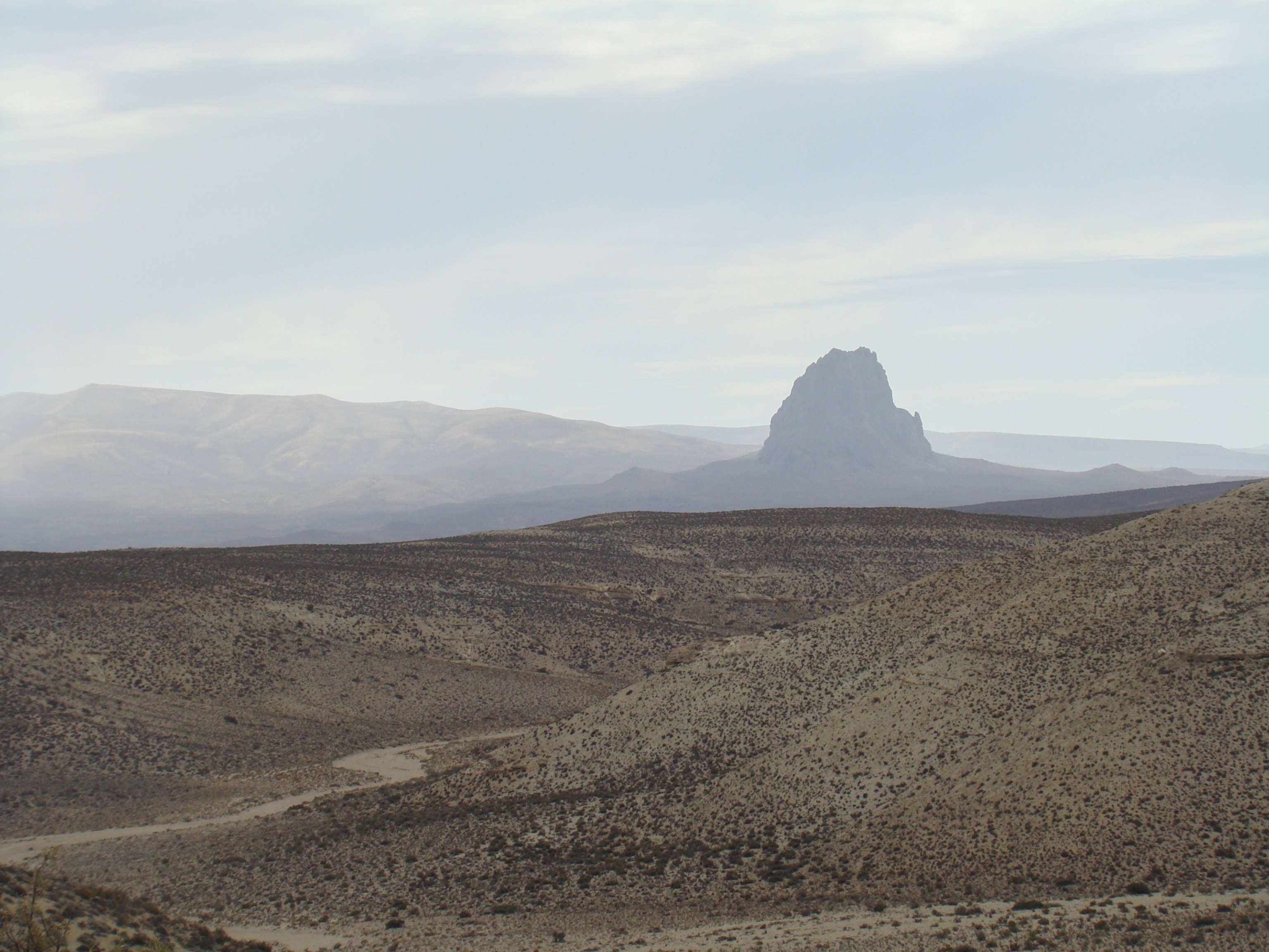 Vast desert landscape with scattered shrubs and undulating hills under a cloudy sky. A prominent rocky formation rises in the distance