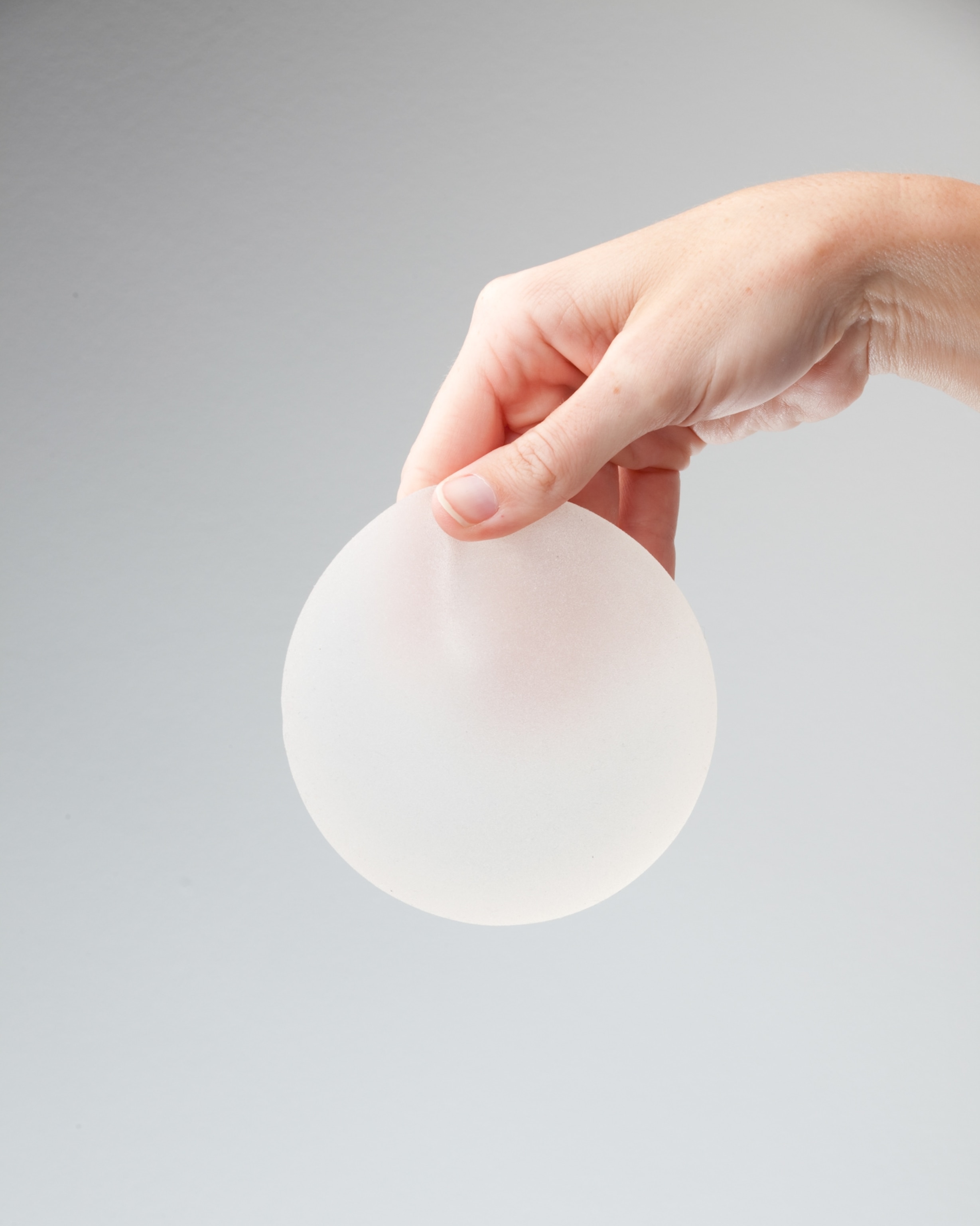 A hand gently holds a round silicone breast implant against a plain gray background