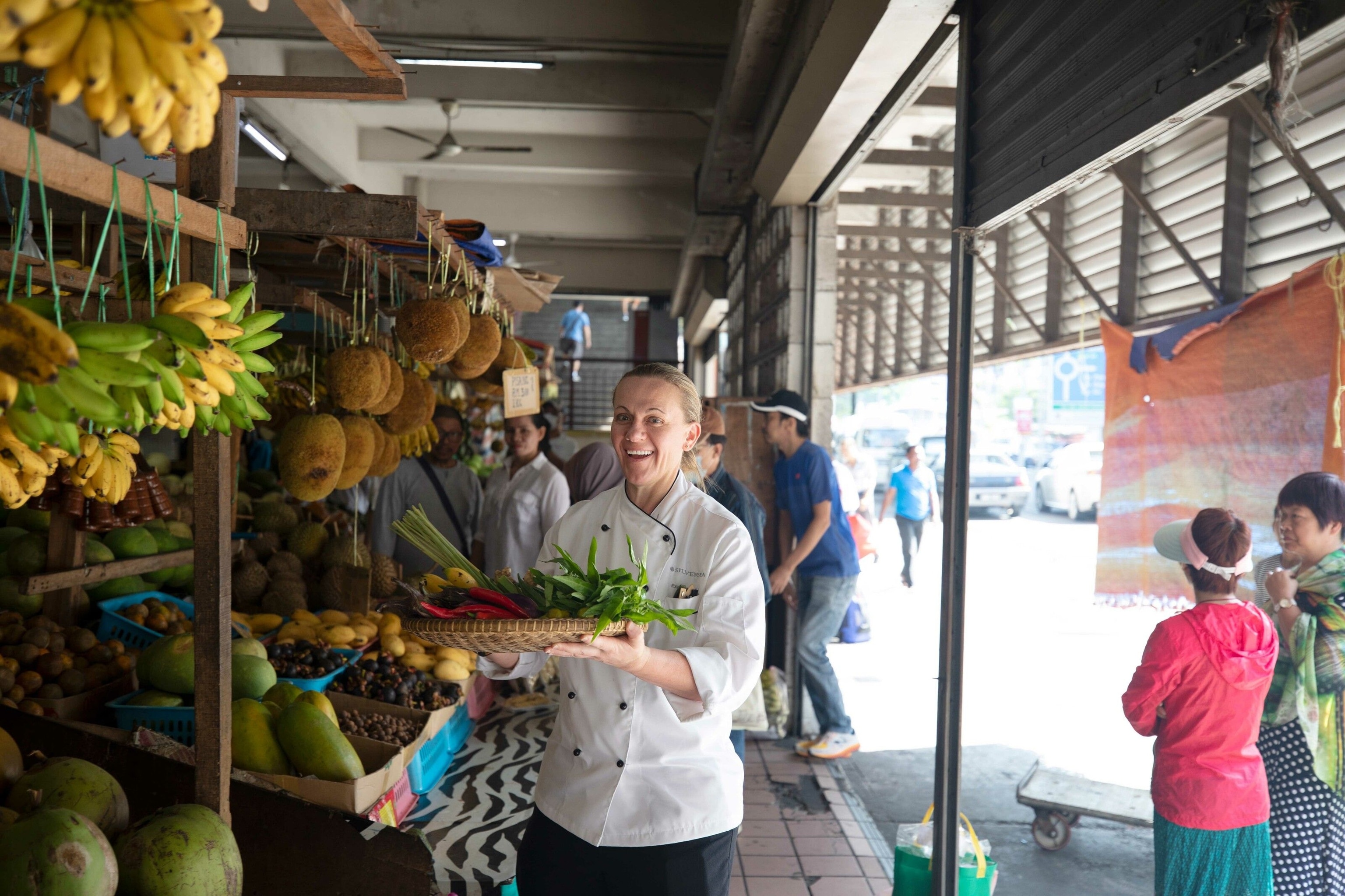 Chef Anne-Mari visits a food market in Vietnam. She wears her white chefs' uniform and carries a woven basket full of green and red vegetables.
