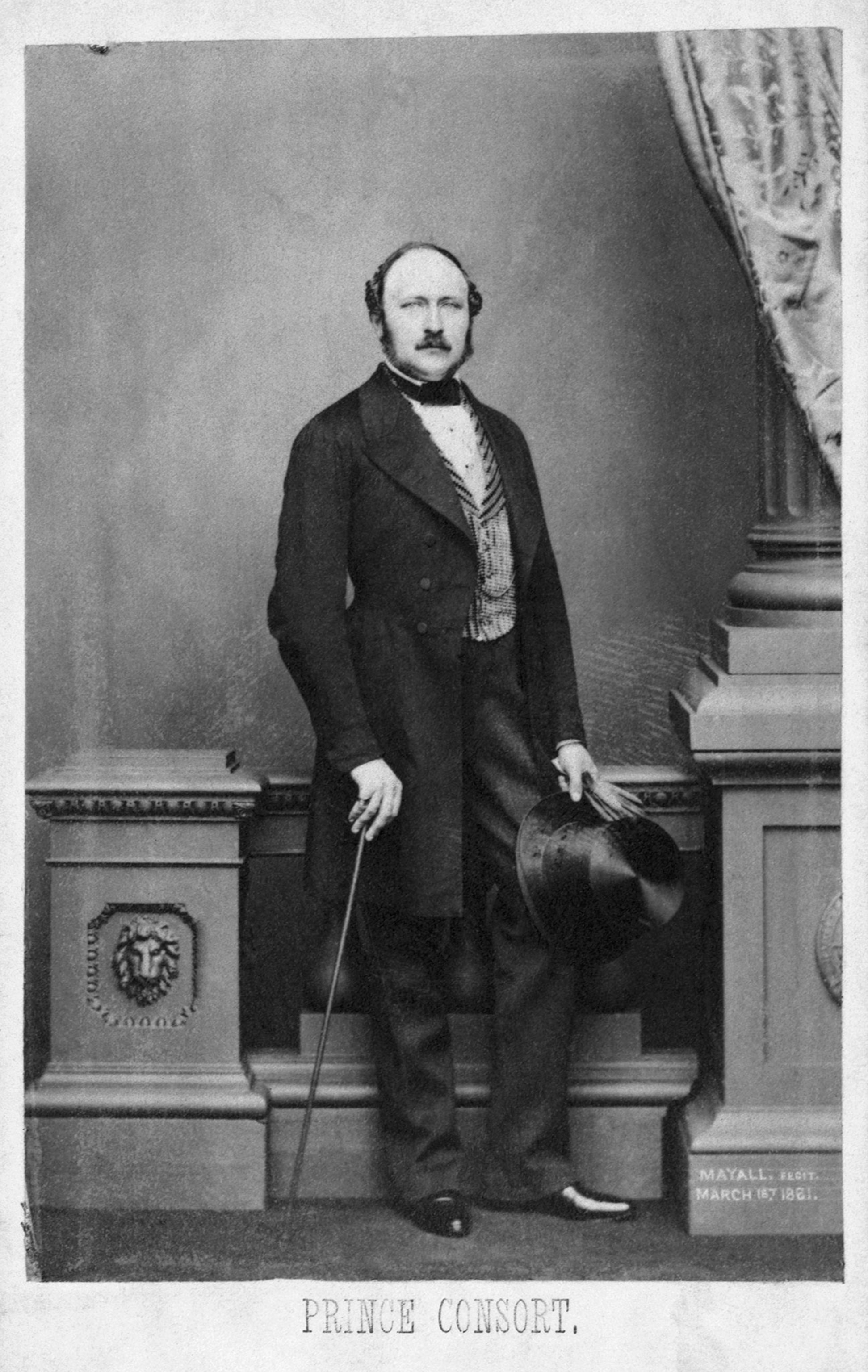 A man holding a cane, top hat and balding, is photographed in black and white.