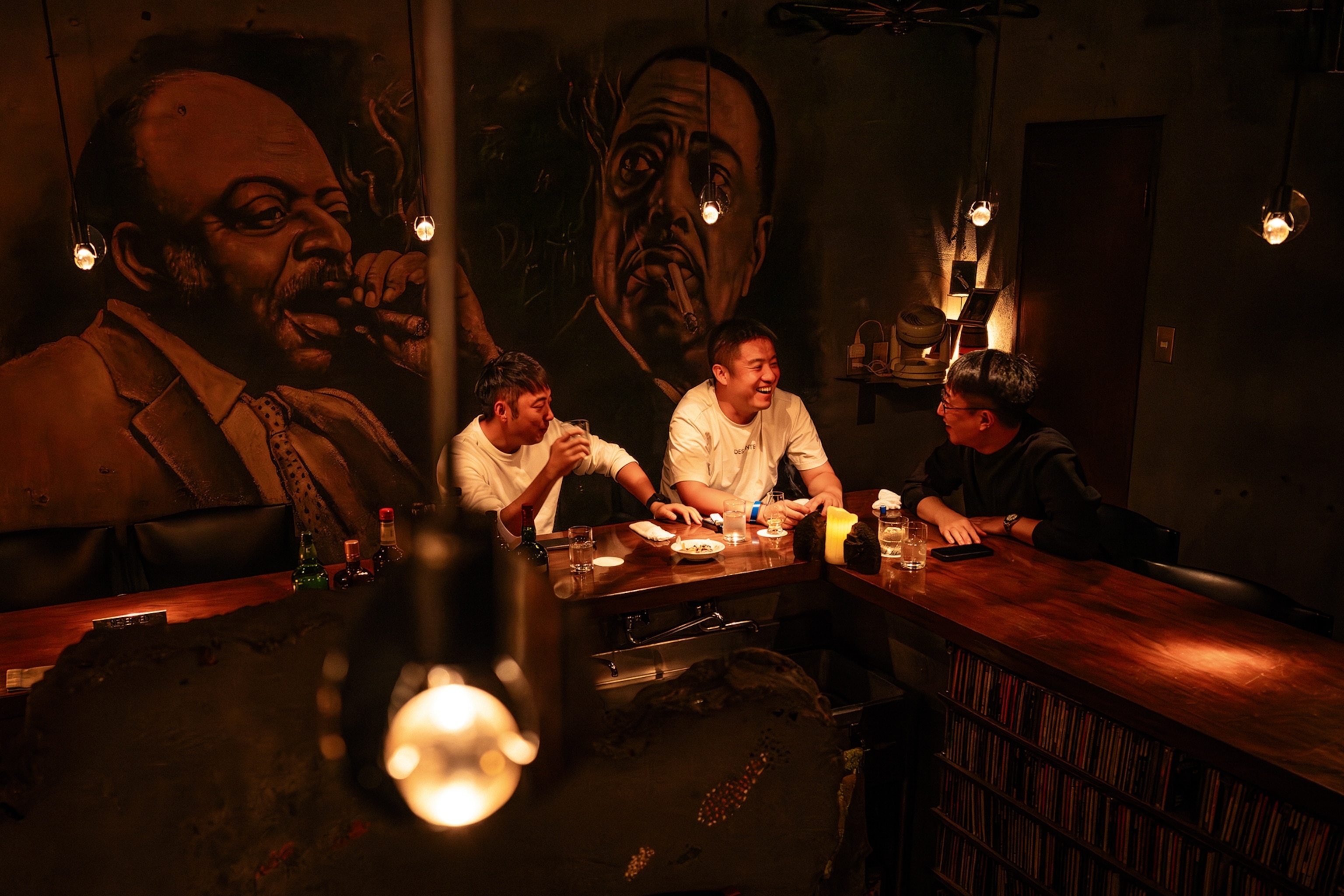 Three local men sitting around the corner of a low-lit bar counter chatting and drinking whiskey.
