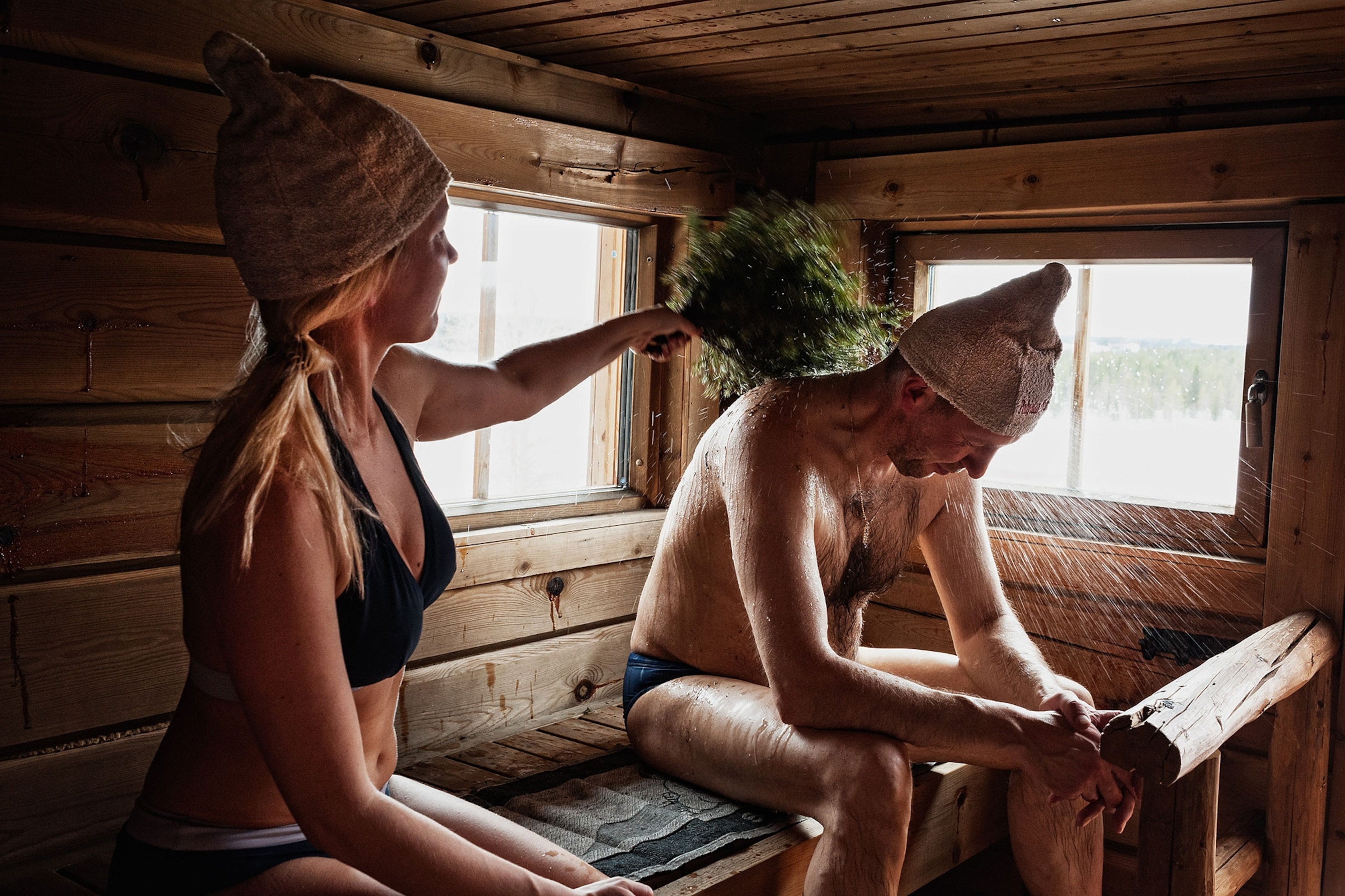 A woman wearing a bikini swimsuit hits a man wearing swimming trunks with a bushel of leaves inside a wooden sauna.