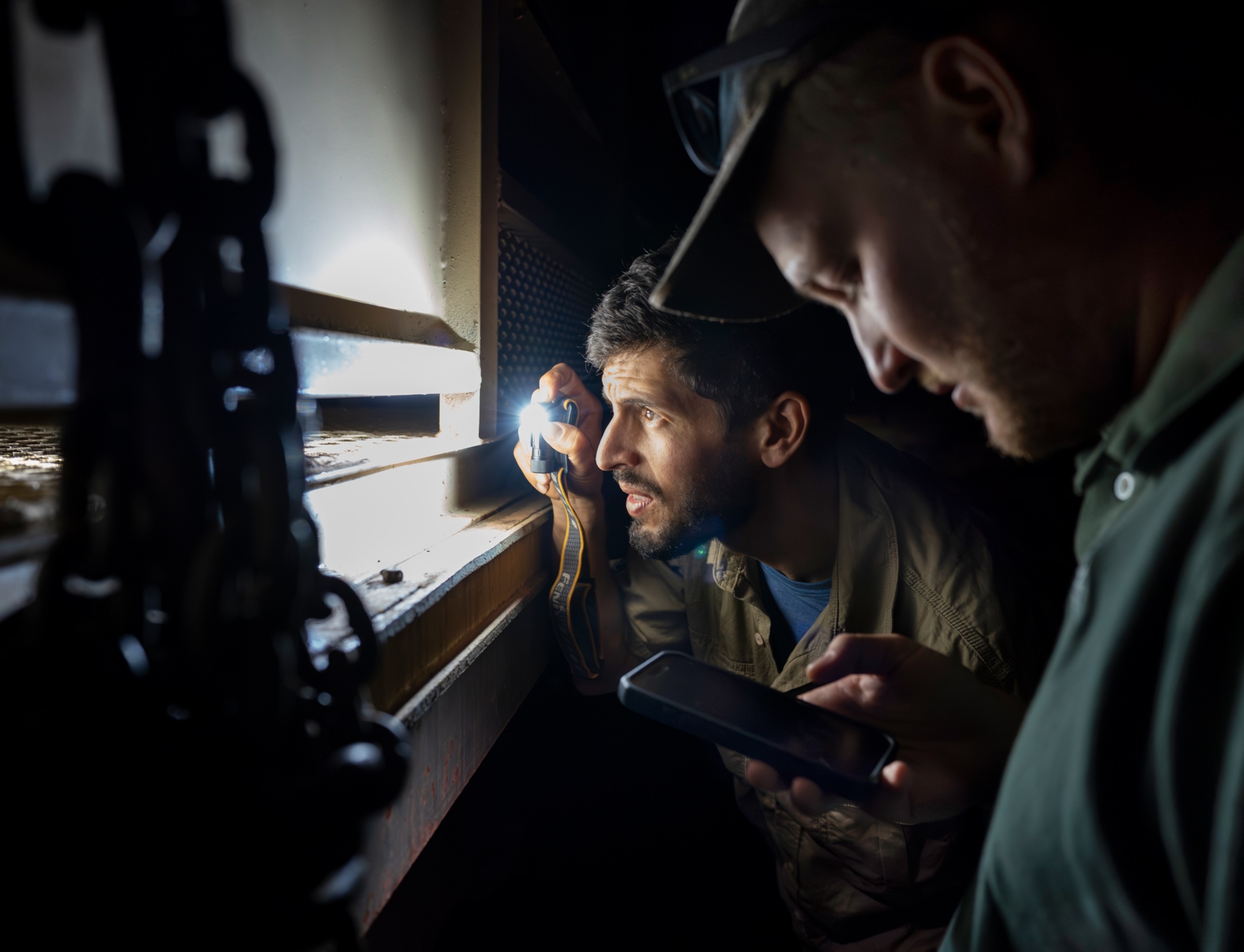 In the dark a man uses a flashlight to look into the bottom of a metal container while another man stands next to him and looks at his phone.