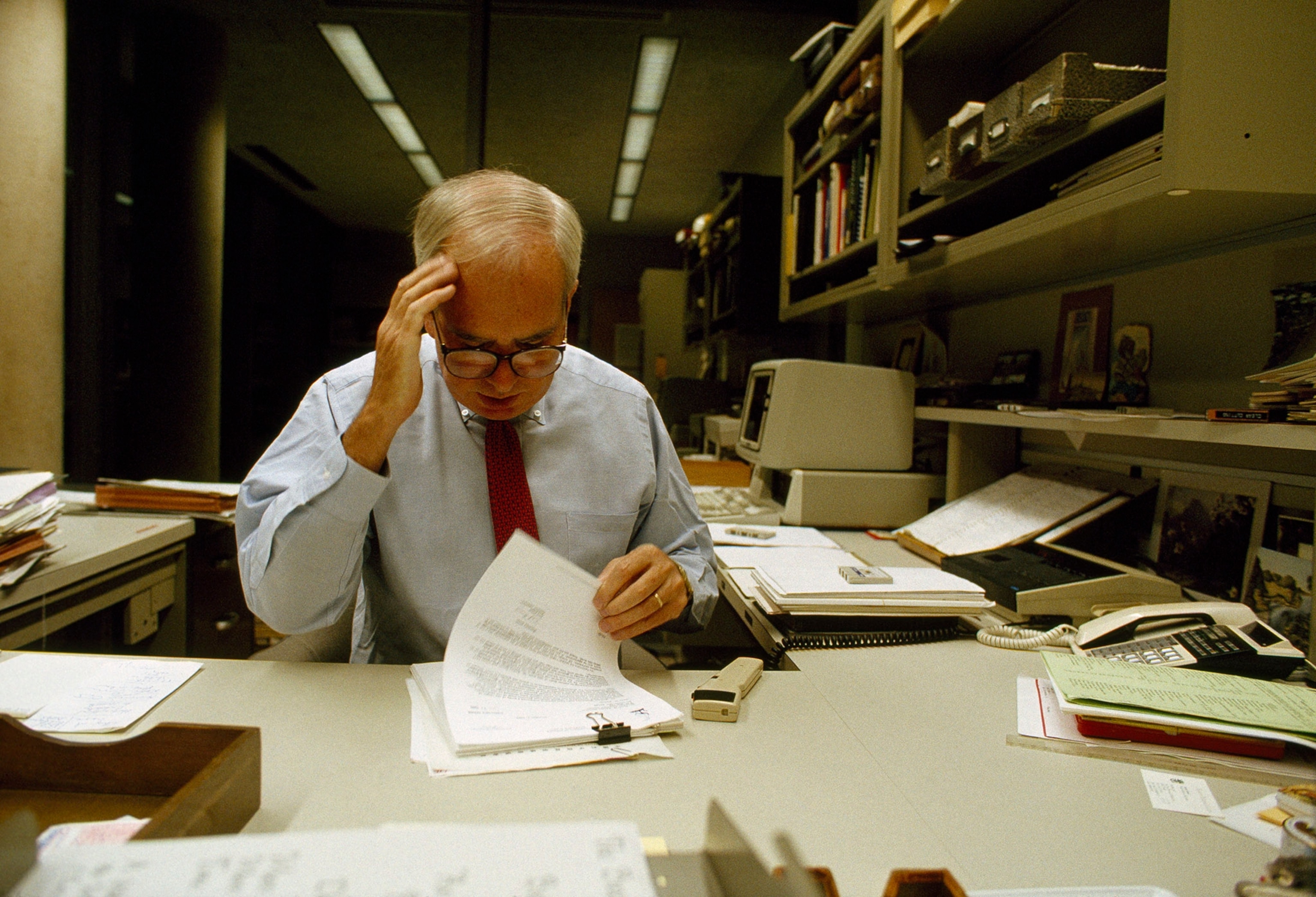 Man in glasses, blue button up shirt, and a red tie sits at a desk looking over papers