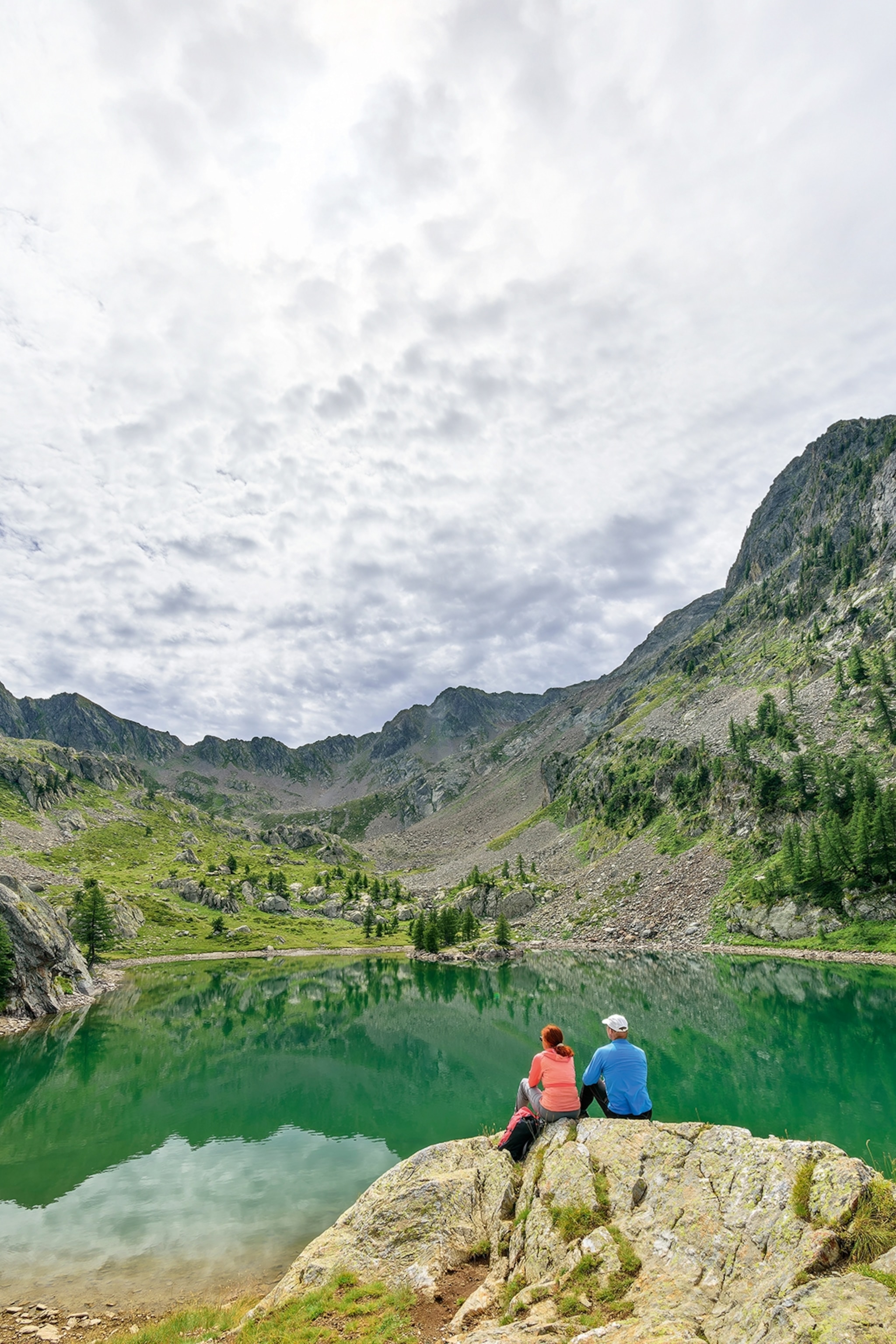 A couple of hikers sitting on the edge of a rock overlooking a clear mountain lake with more mountains in the background.