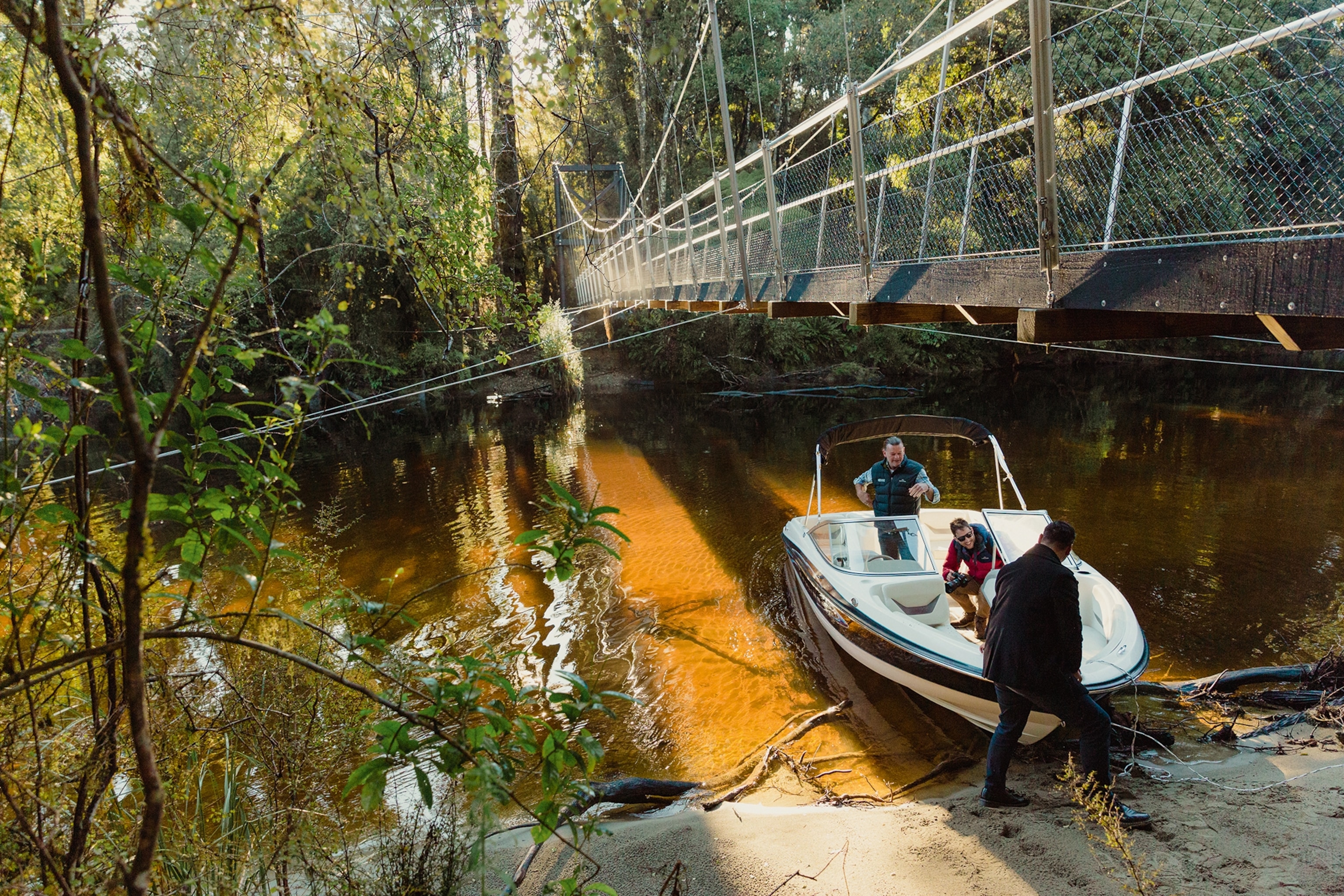 A wide shot taken at eye-level to a swing bridge, hanging low over a forest river with a manned boot anquored on the river bank.