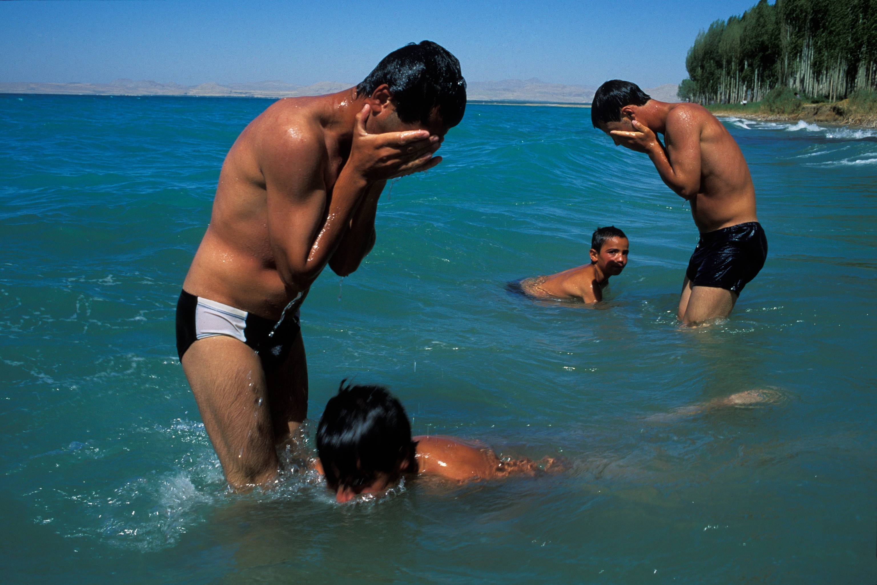 Two men standing covering their face and two younger boys in the water swimming.
