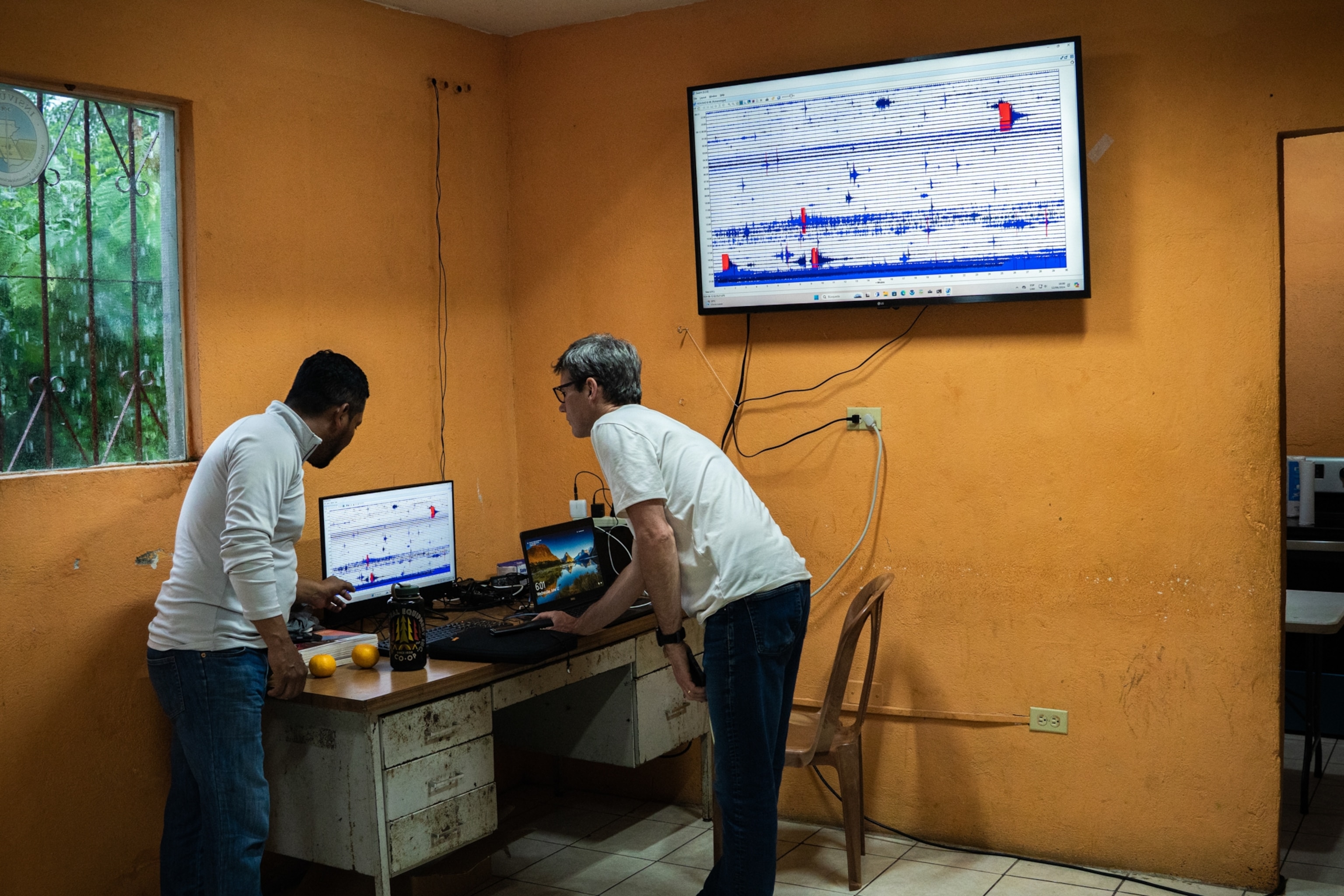 Two people analyzing data in a room with orange walls. A large monitor displays graphs, and a laptop shows landscape imagery
