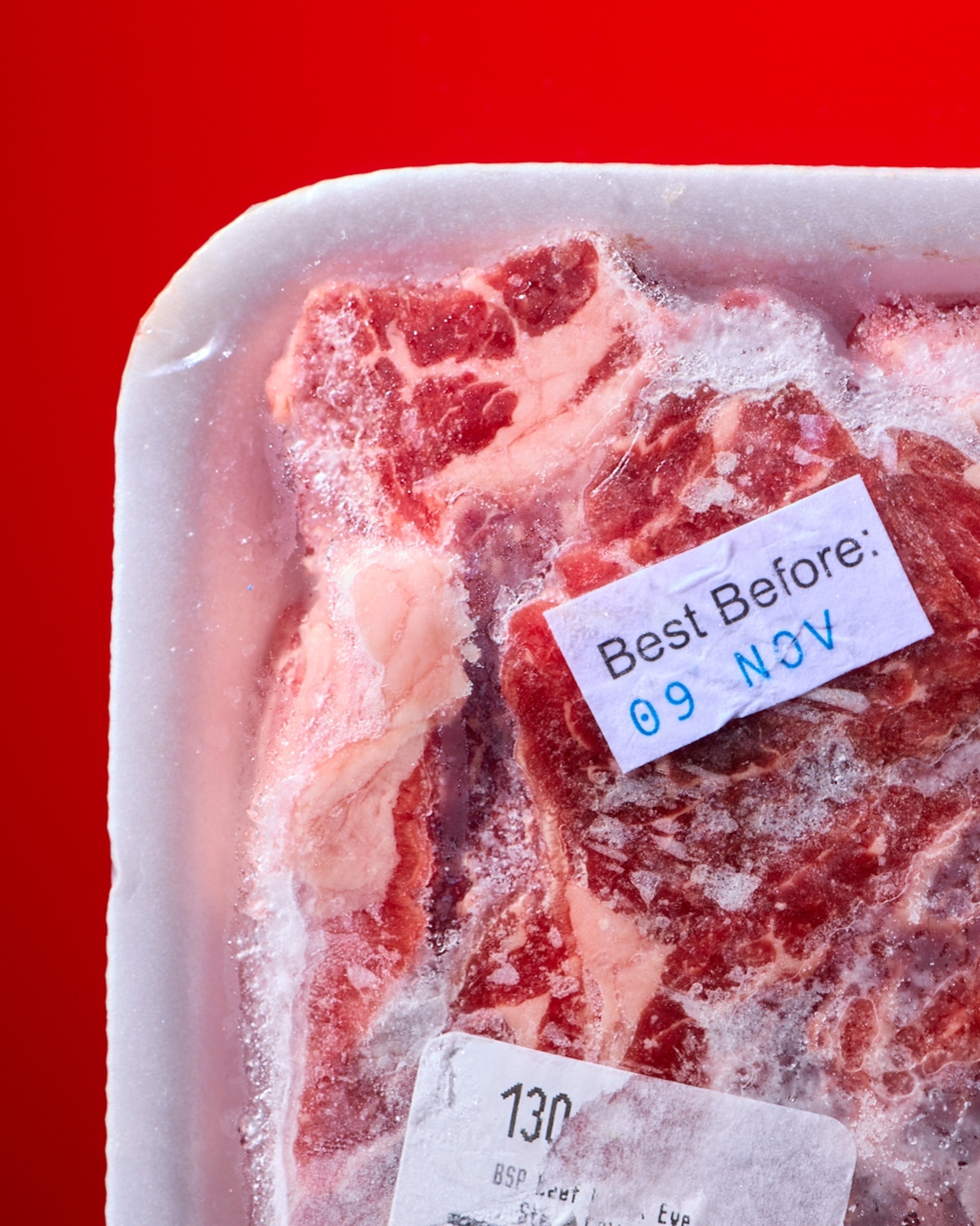 Frozen meat in a white tray against a red background, labeled "Best Before: 09 NOV" with frost visible