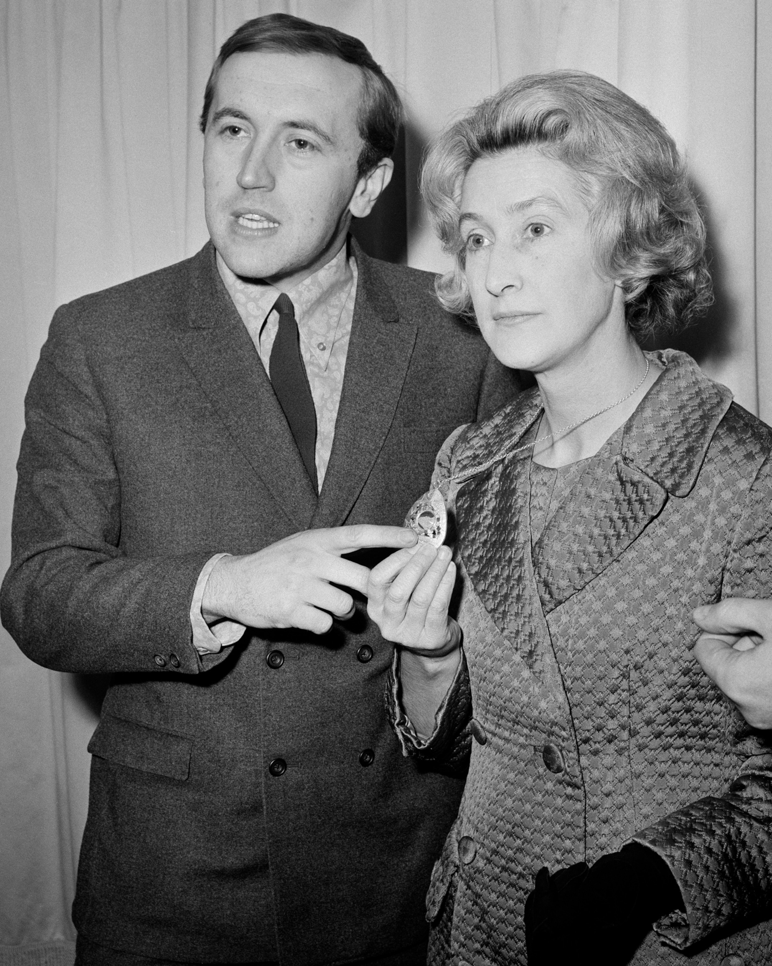 A man stands next to a woman and points to the necklace that she is wearing.